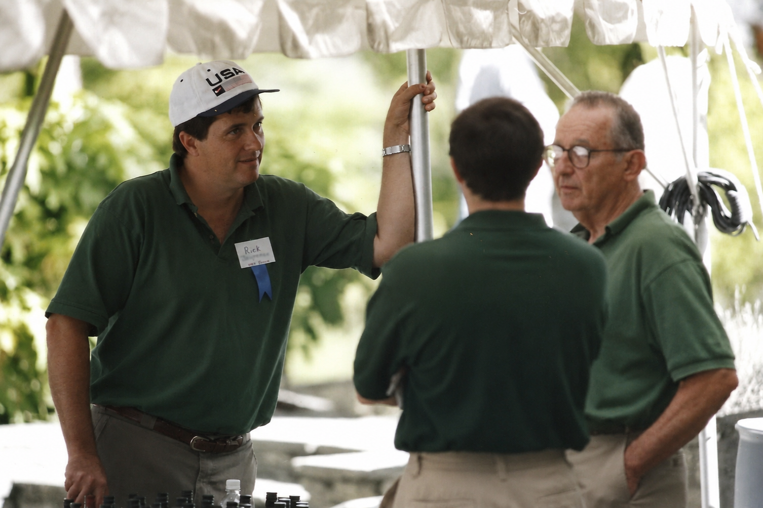 Three men, two with dark hair and one with gray hair and glasses, are engaged in conversation under a tent. They are wearing green shirts, and one has a white cap with a USA logo. The man with the cap is named Rick, as indicated by his name tag.