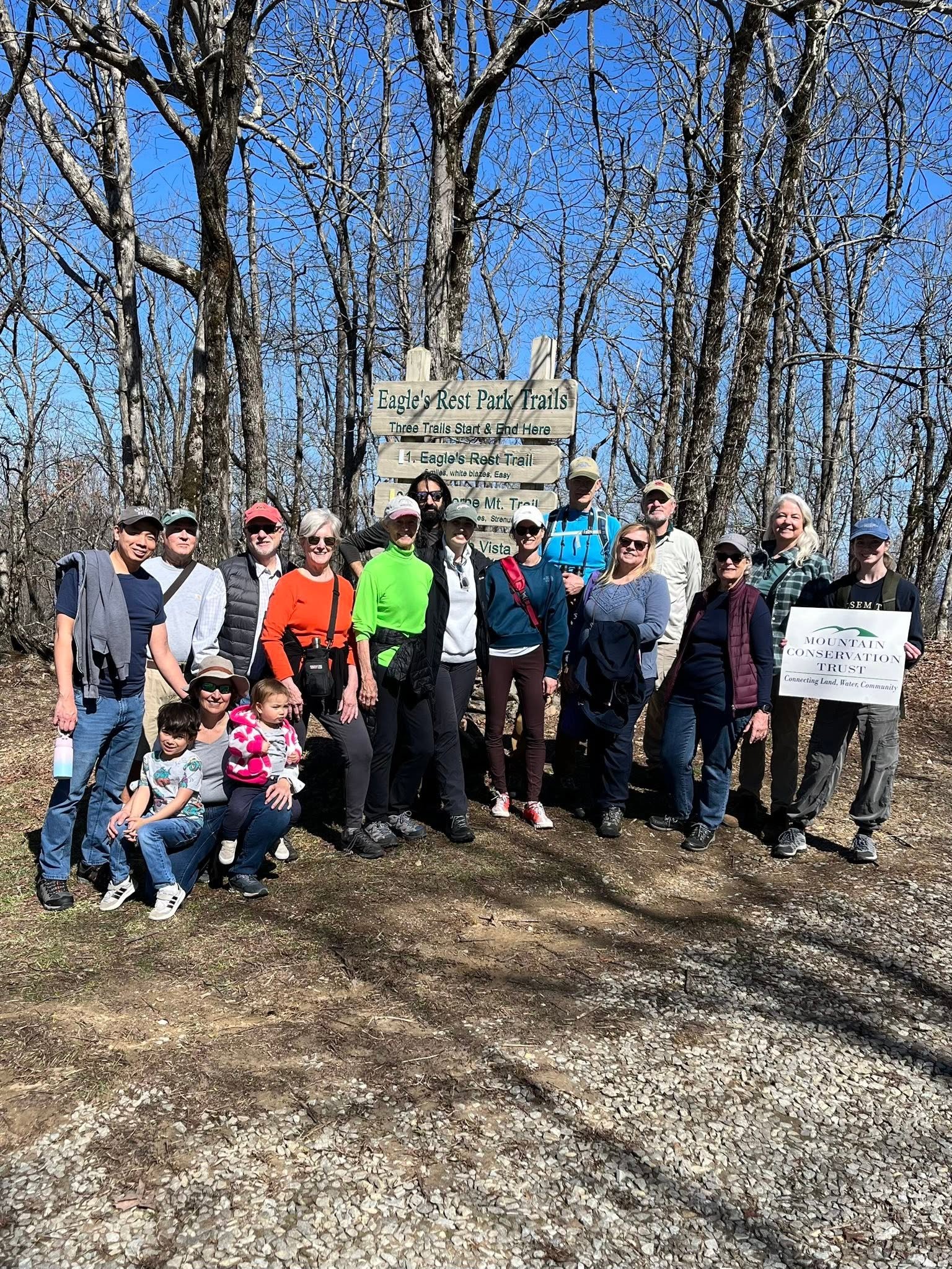 A group of people posing outdoors on a trail in a wooded area with leafless trees, a blue sky, and a sign that reads 'Eagle's Rest Park Trails'.