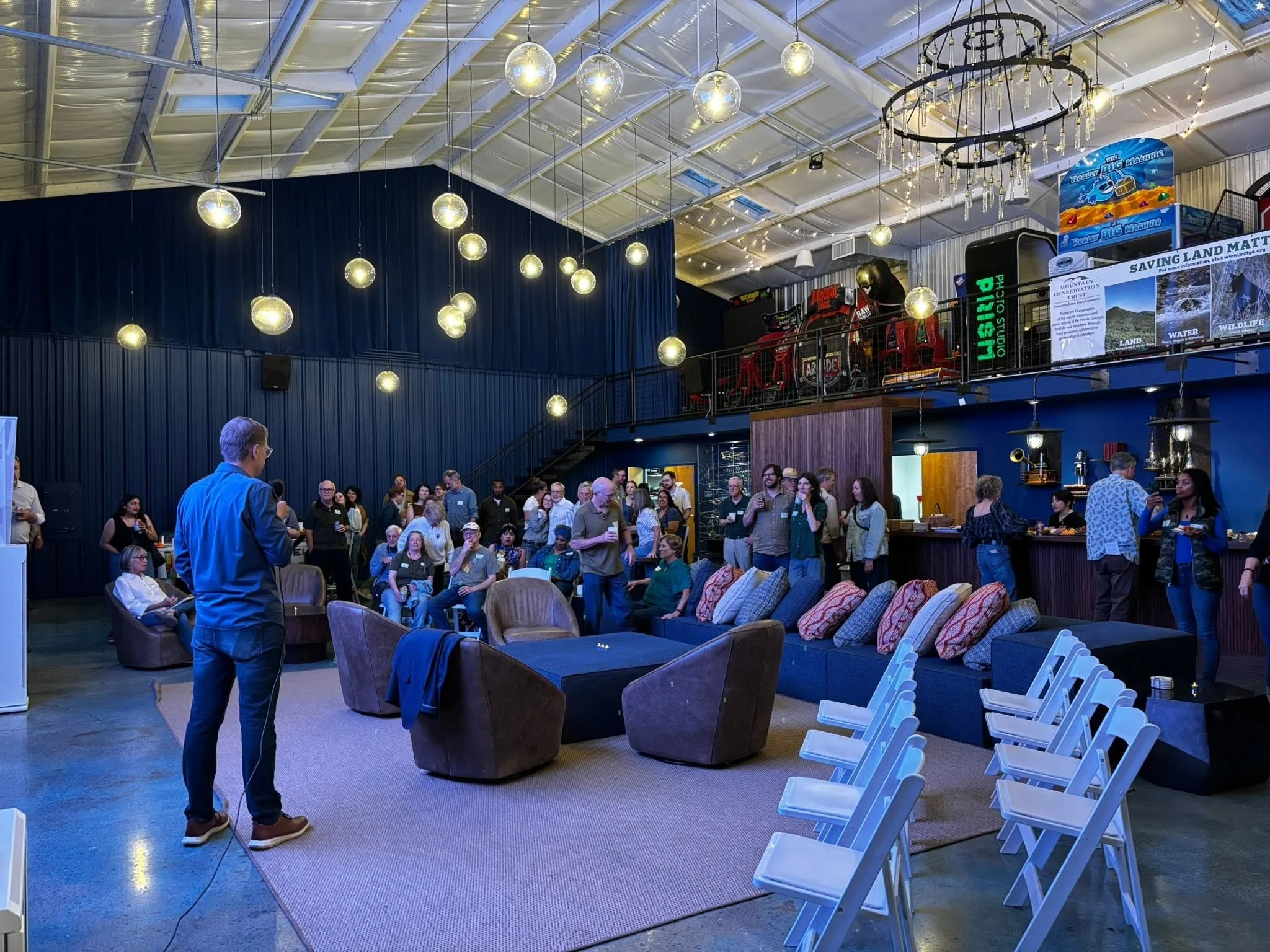 An indoor event space with a large crowd of people standing and sitting, decorated with hanging round light fixtures and string lights, with a mezzanine level containing arcade games and promotional displays.