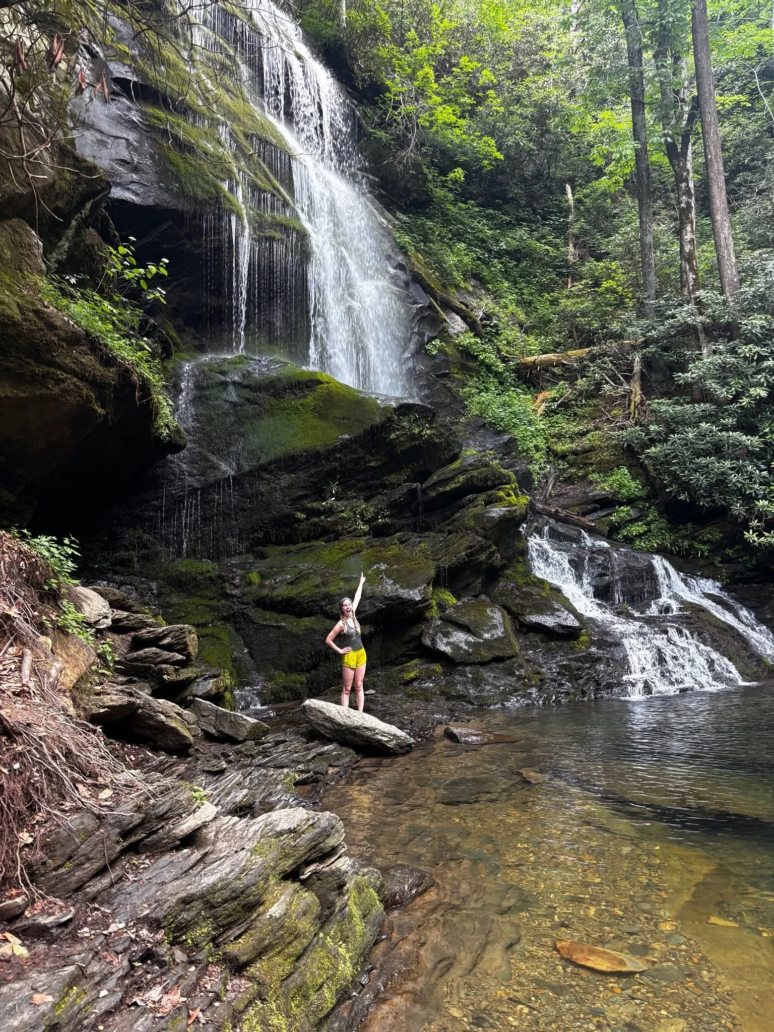 A person stands on rocks in a shallow stream at the base of a small waterfall surrounded by green trees and moss-covered rocks.