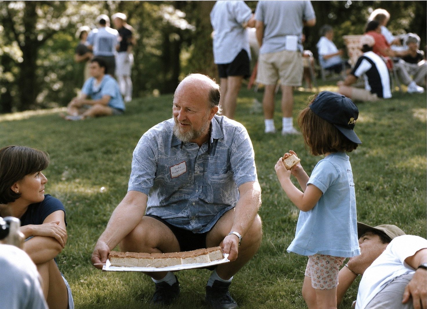 A group of people, including an older man with a name tag, sitting on the grass at a park, with a young girl holding a slice of cake, while others stand and sit in the background.