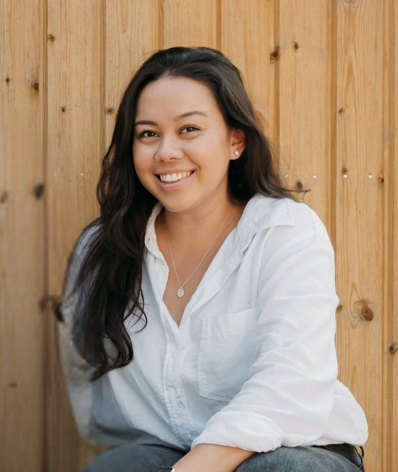 A woman with long dark hair smiling, wearing a white shirt, silver jewelry, and sitting in front of a wooden fence.
