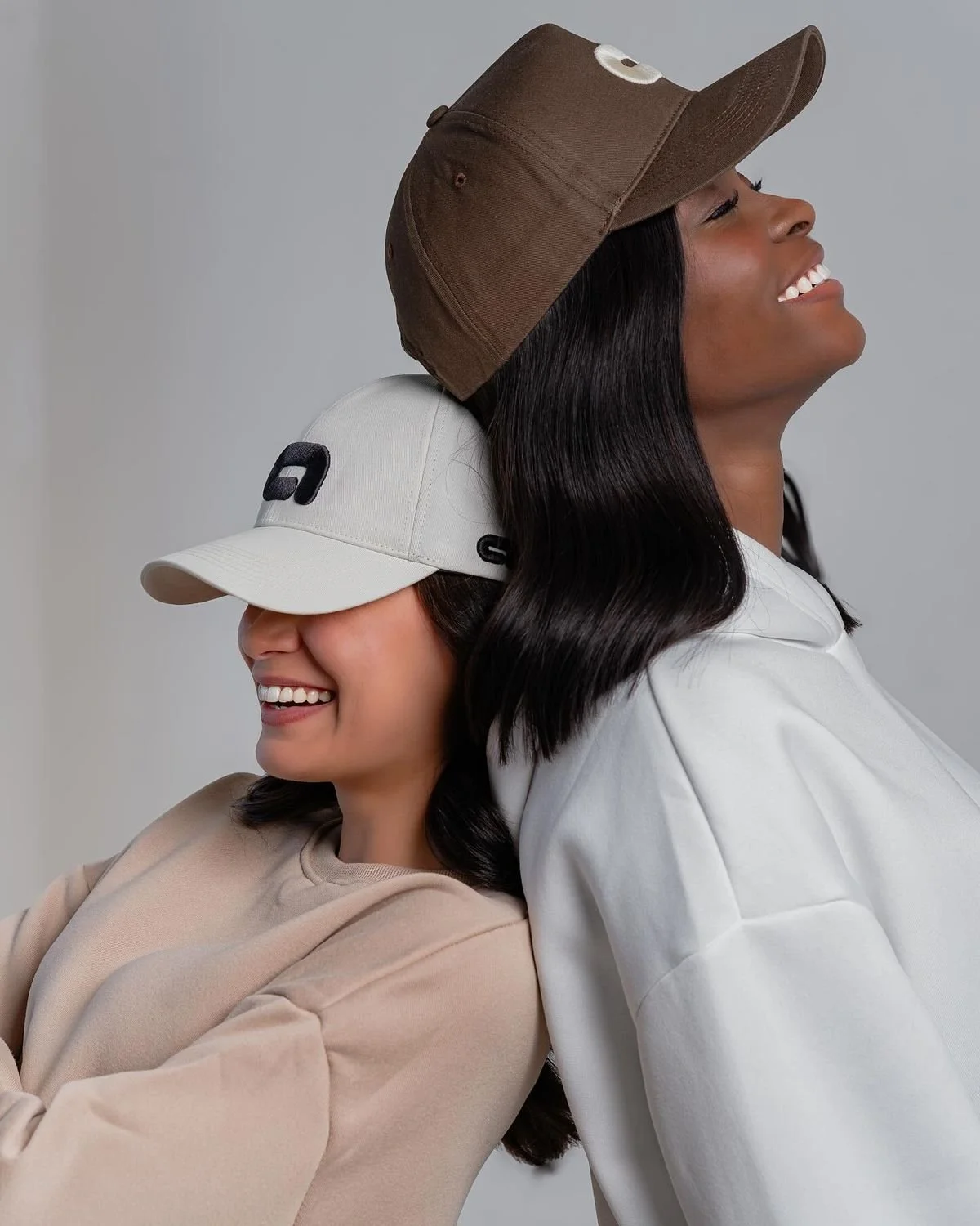 Two women smiling and leaning on each other, wearing baseball caps, one beige and one white, against a gray background.