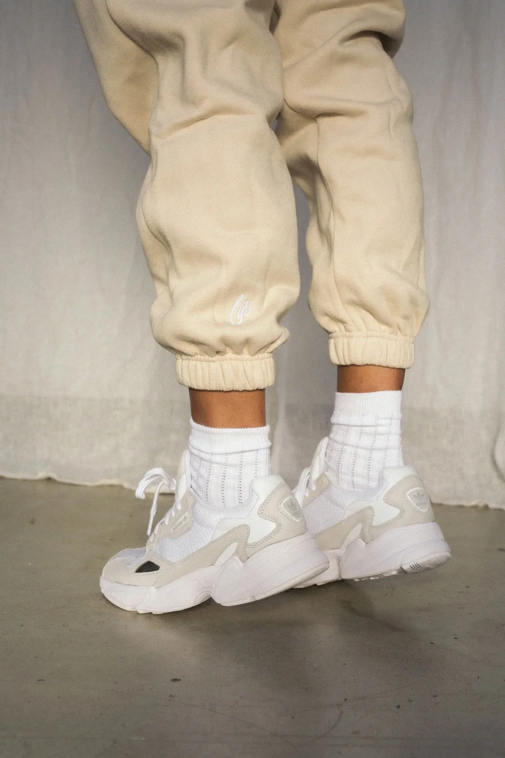 Close-up of a person's legs wearing beige jogger pants, white socks, and white athletic sneakers standing on a gray floor against a neutral background.