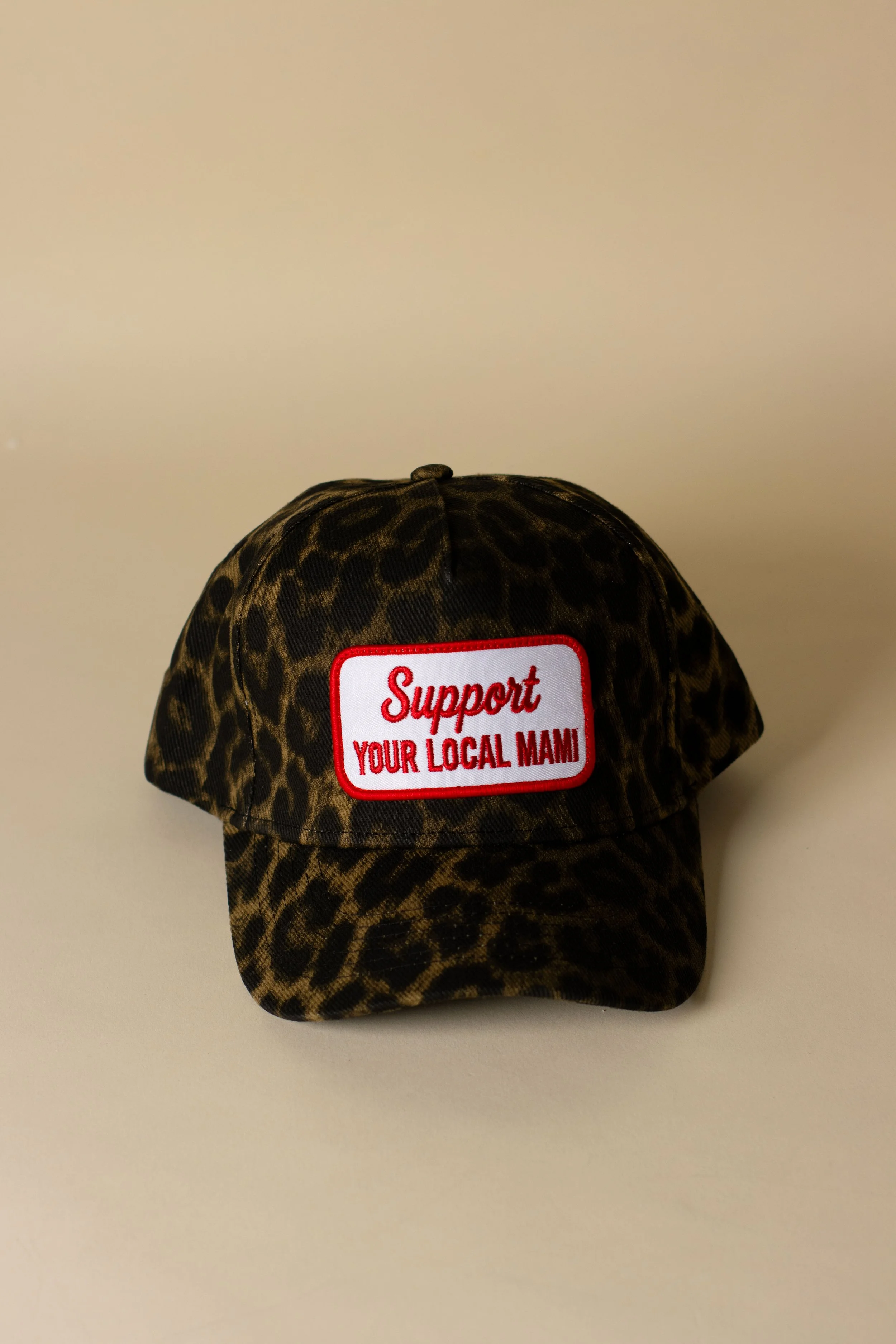 A leopard-print cap with a red and white support patch that says 'Support Your Local Mami' against a plain beige background.