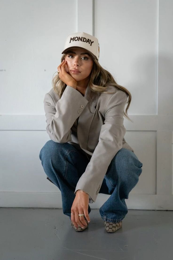 A woman in a beige blazer and jeans squatting against a white wall, wearing a beige cap with 'MONDAY' written on it, resting her face on her hand with a thoughtful expression.