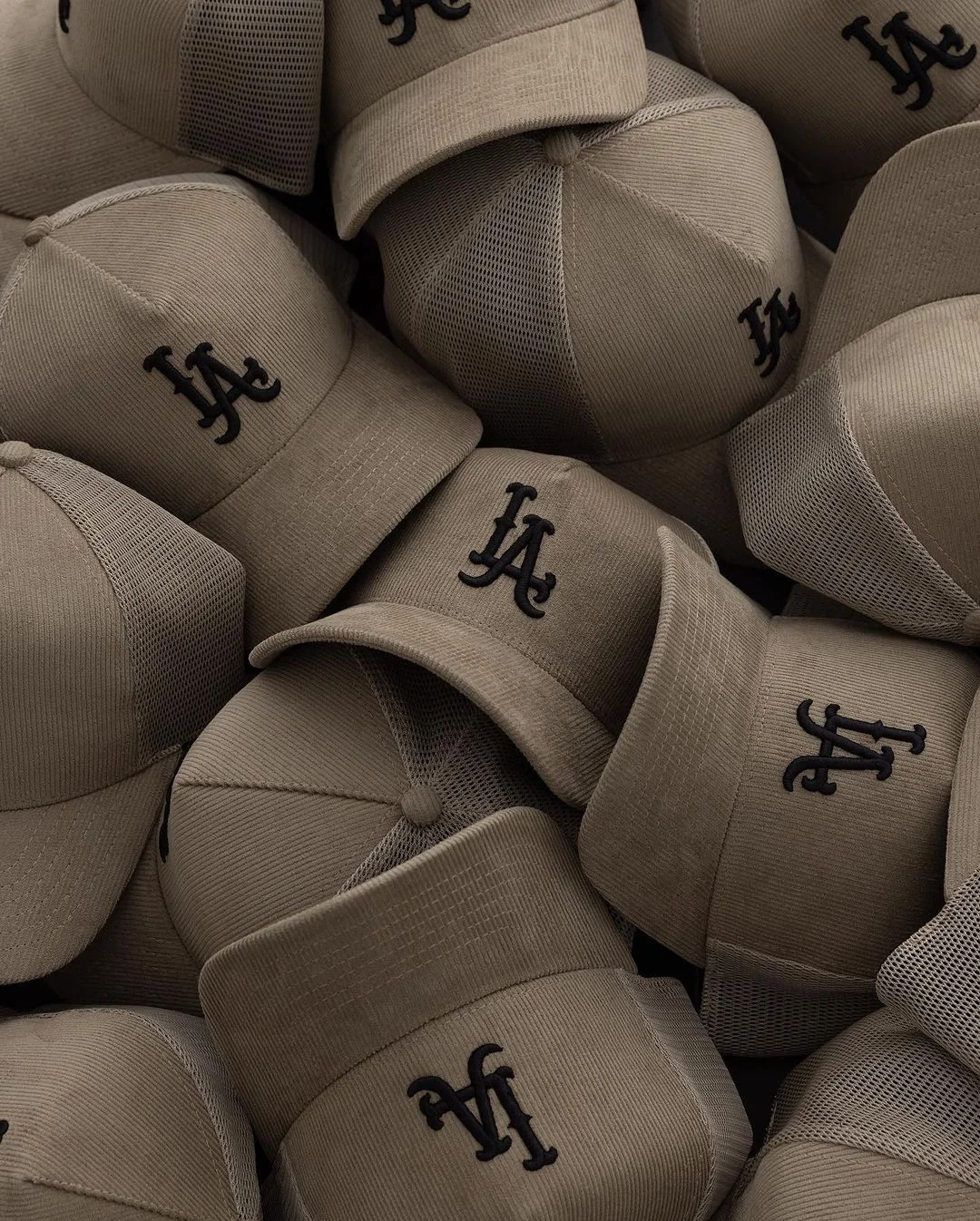 A pile of beige baseball caps with black embroidered 'LA' logos, some caps are facing forward and others are angled.