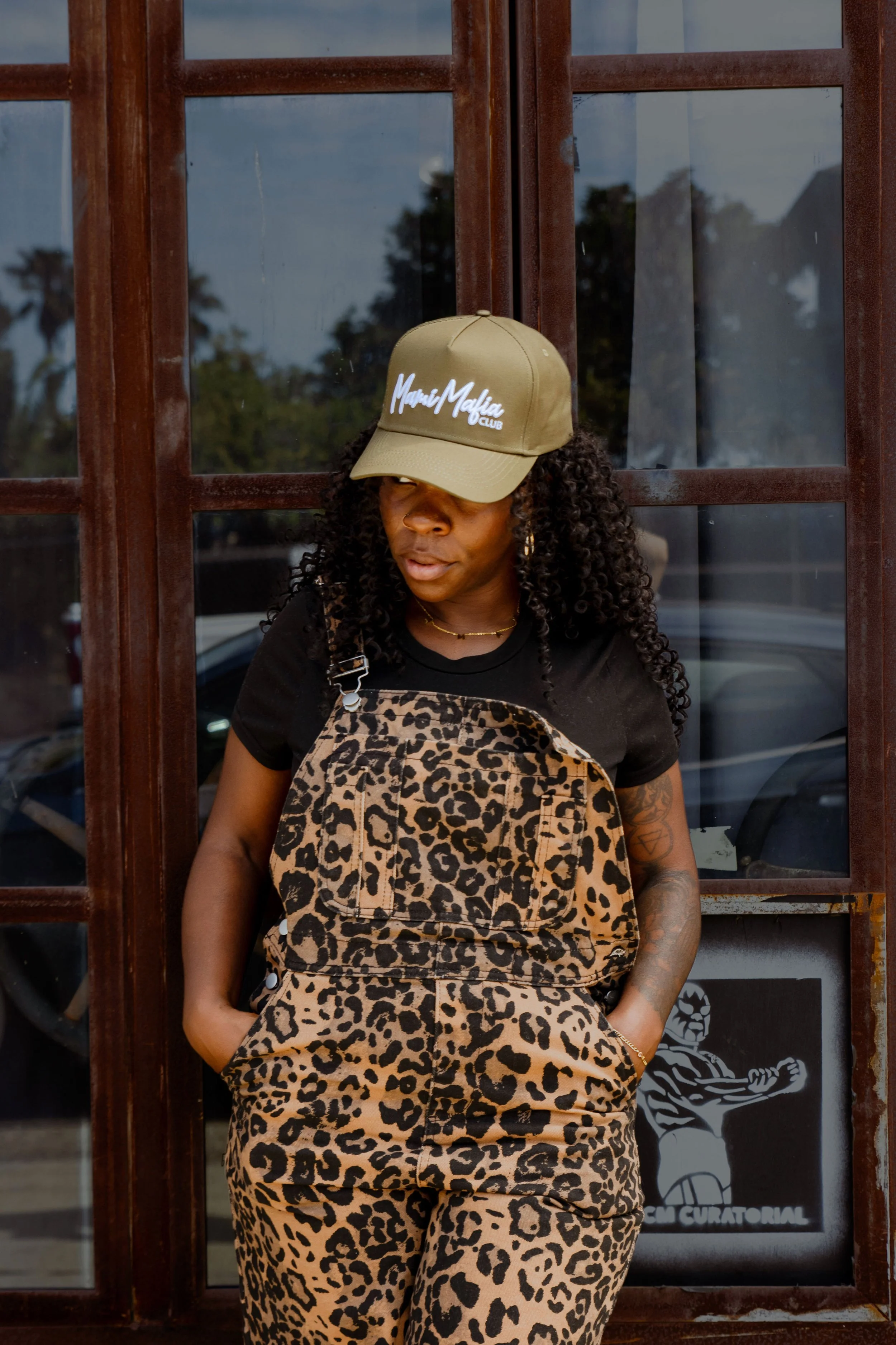 A woman with curly hair wearing a baseball cap that says "Mafia Mafia Club," a black T-shirt, and leopard print overalls, standing in front of a wooden window with glass panes.