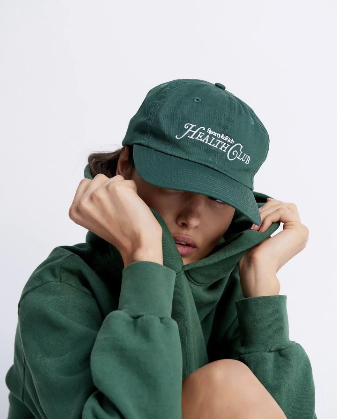 Person wearing a green cap with the words "Sporty & Rich HEALTH CLUB" embroidered on it, and a matching green hoodie, resting their head on their hand.