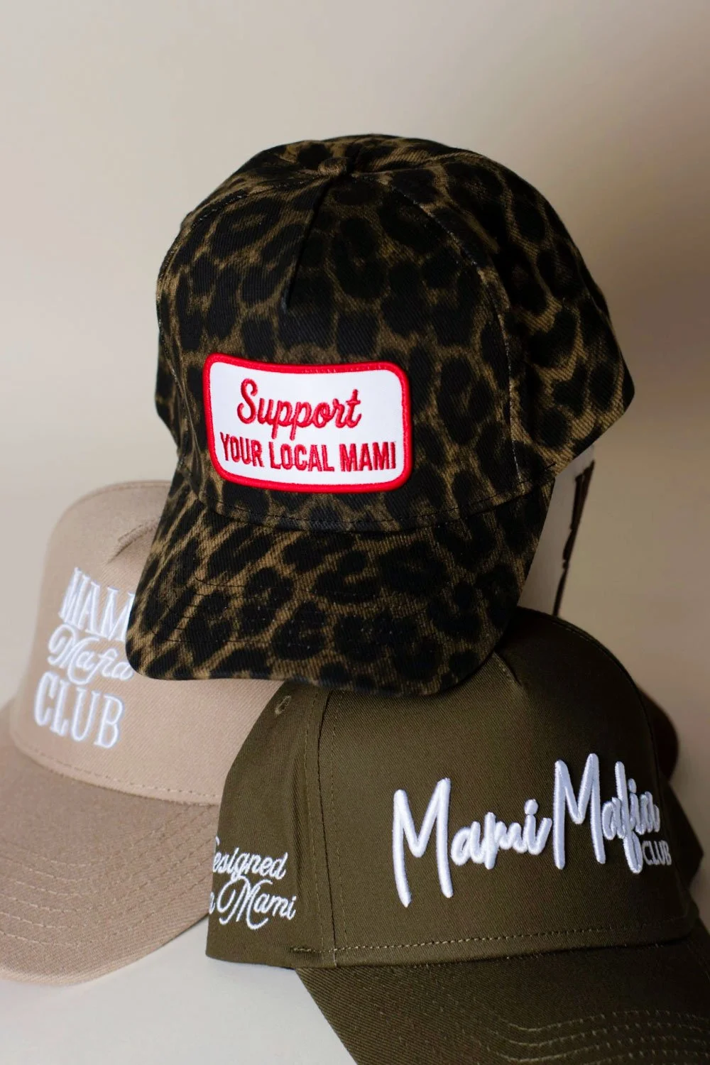 Three baseball caps, one with a leopard print pattern and a red and white patch that says support your local MAMI, and two plain caps with embroidered text supporting the Mami Mafia club.