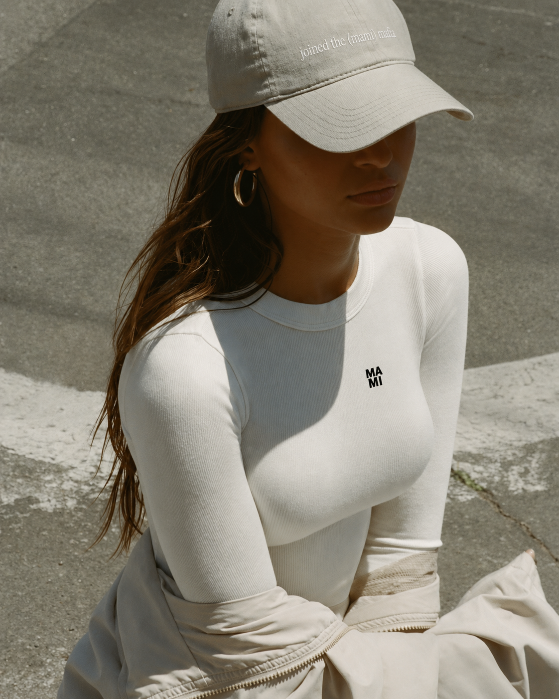 A woman sitting on the street wearing a beige baseball cap with 'joined the (mami) mafia' embroidered, a white long-sleeve shirt with 'MAMI' printed on the chest, and large hoop earrings.