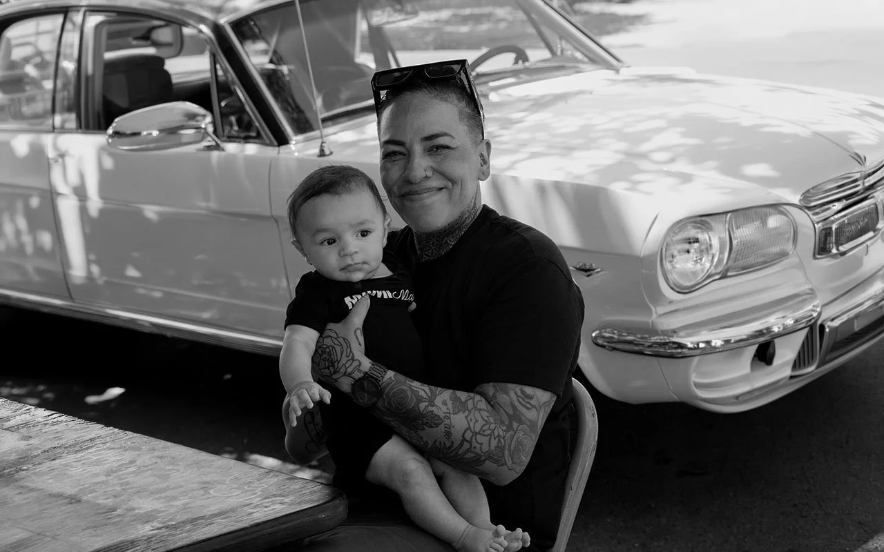 A woman with tattoos holding a baby in front of a vintage car