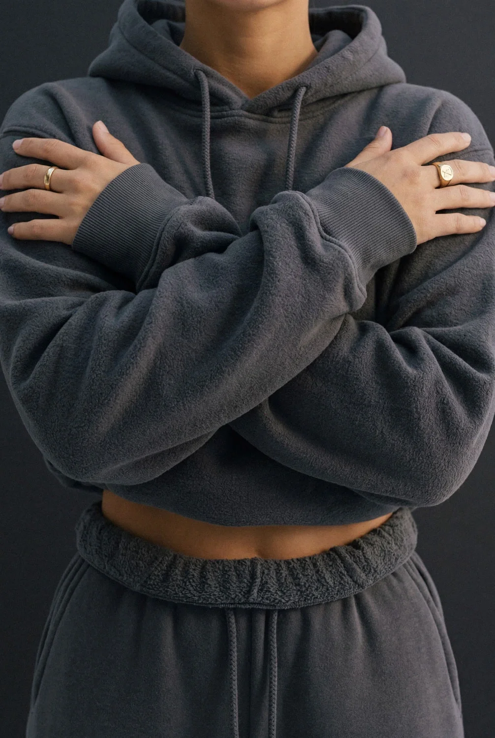Person standing with arms crossed, wearing a gray hoodie and sweatpants, with rings on fingers, against a dark background.