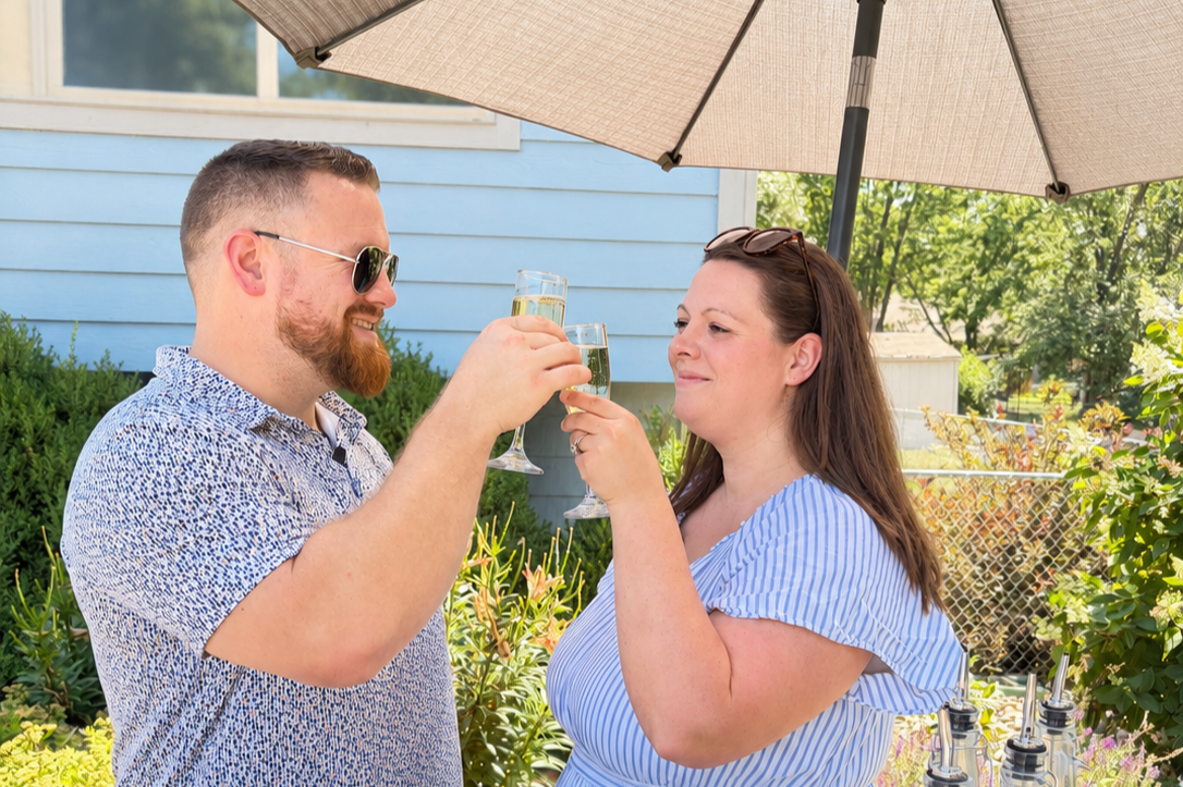 A man and a woman clinking glasses of champagne outdoors under an umbrella, with greenery and a blue house in the background.
