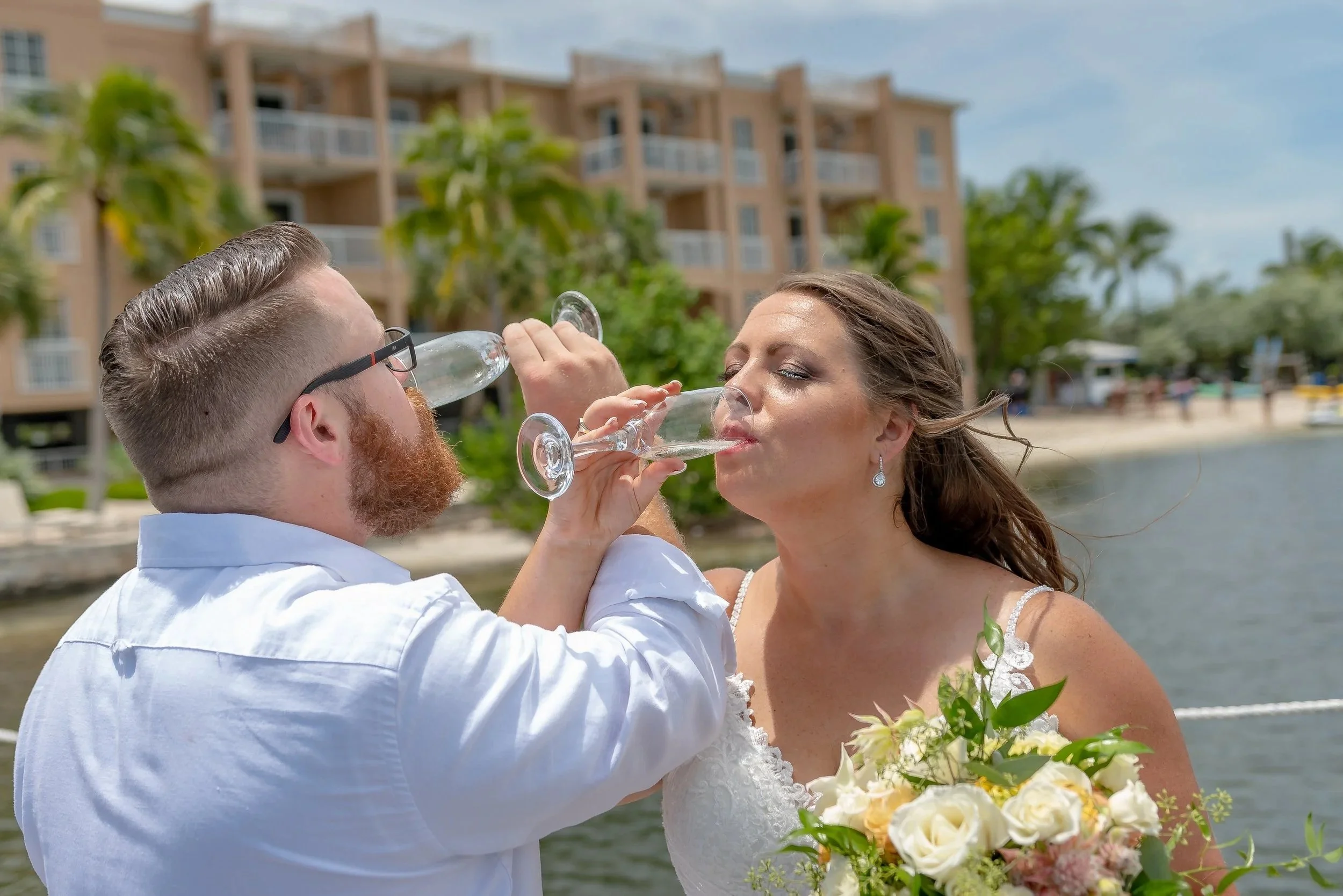 A man and woman in wedding attire sharing a drink at an outdoor waterfront wedding.