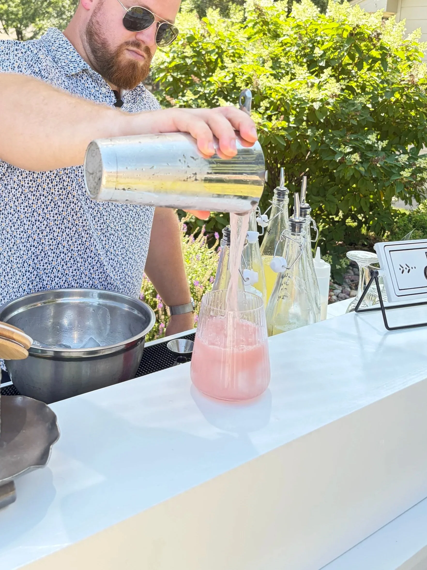 A man with sunglasses and a beard pouring light pink drink from a cocktail shaker into a glass with the same drink. Bar setup with bottles and accessories outdoors on a sunny day.