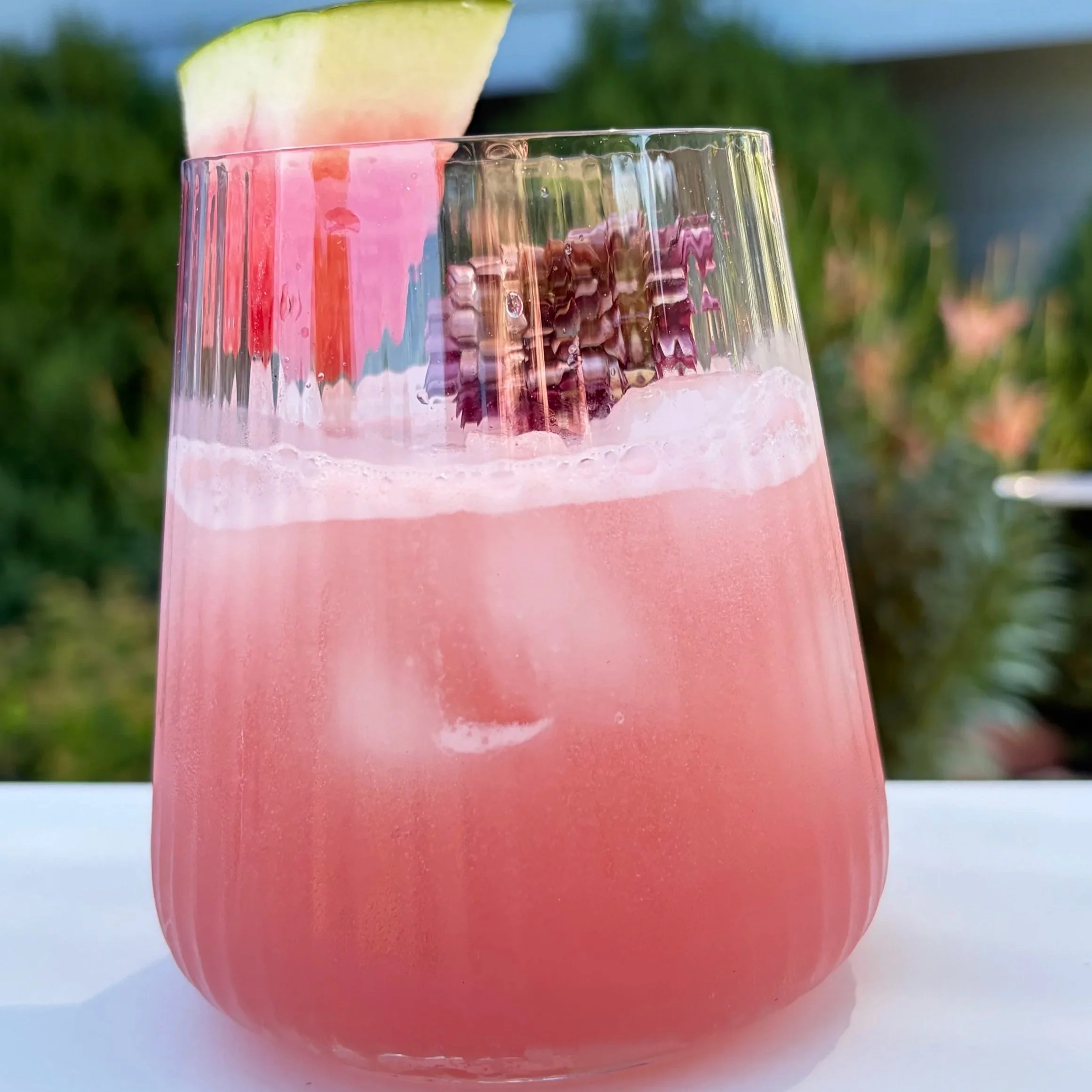 Close-up of a pink-colored drink in a transparent glass with an apple slice garnish, set outdoors with a blurred green background.