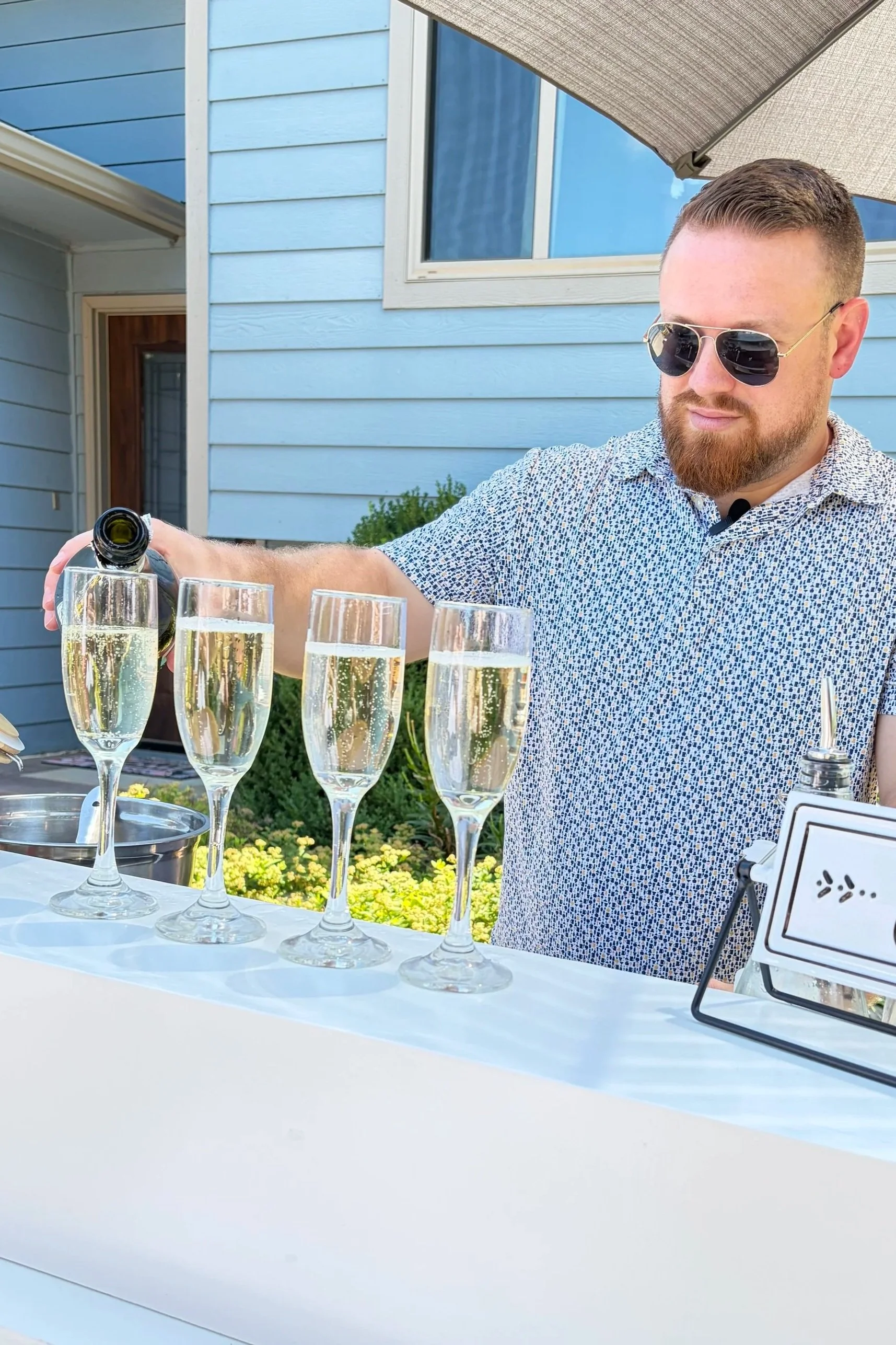 A man wearing sunglasses and a patterned shirt pours champagne into flutes on a table set for a celebration outdoors, with a light blue house and a patio umbrella in the background.