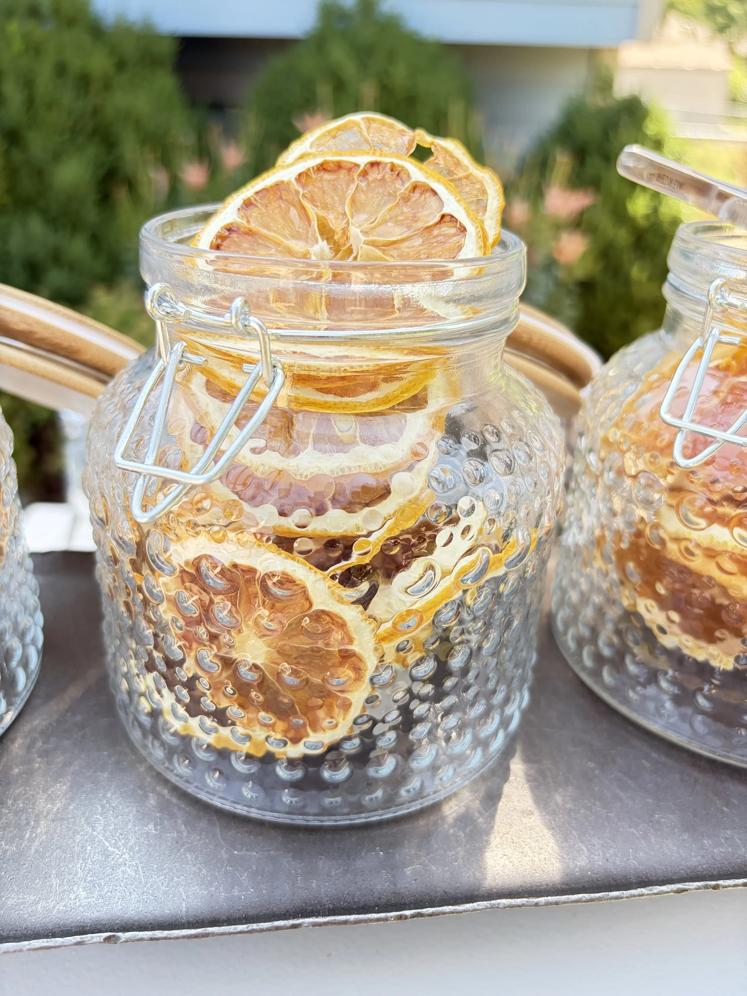 A glass jar with a textured surface filled with dried orange slices. Additional jars with similar contents are partially visible around it, on a metallic surface outdoors with greenery in the background.
