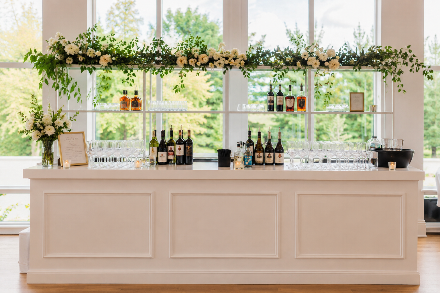 Empty bar with white counter, bottles of alcohol, glasses, white flowers in vases, and a floral arrangement on a shelf behind, with large windows showing green trees outside.
