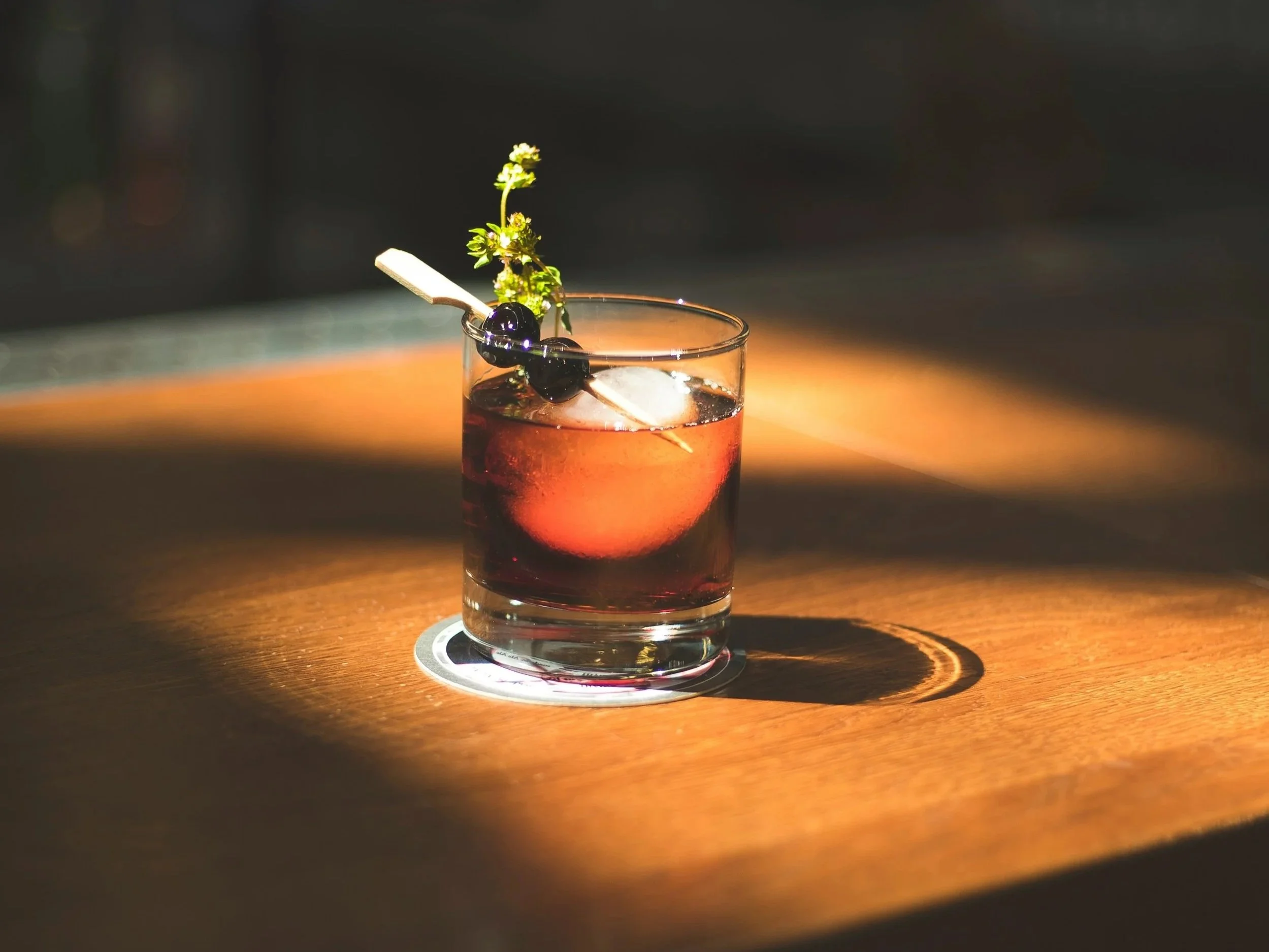 A glass of dark-colored beverage garnished with a sprig of fresh herbs, blueberries, and a small wooden stick, placed on a wooden surface with warm lighting.