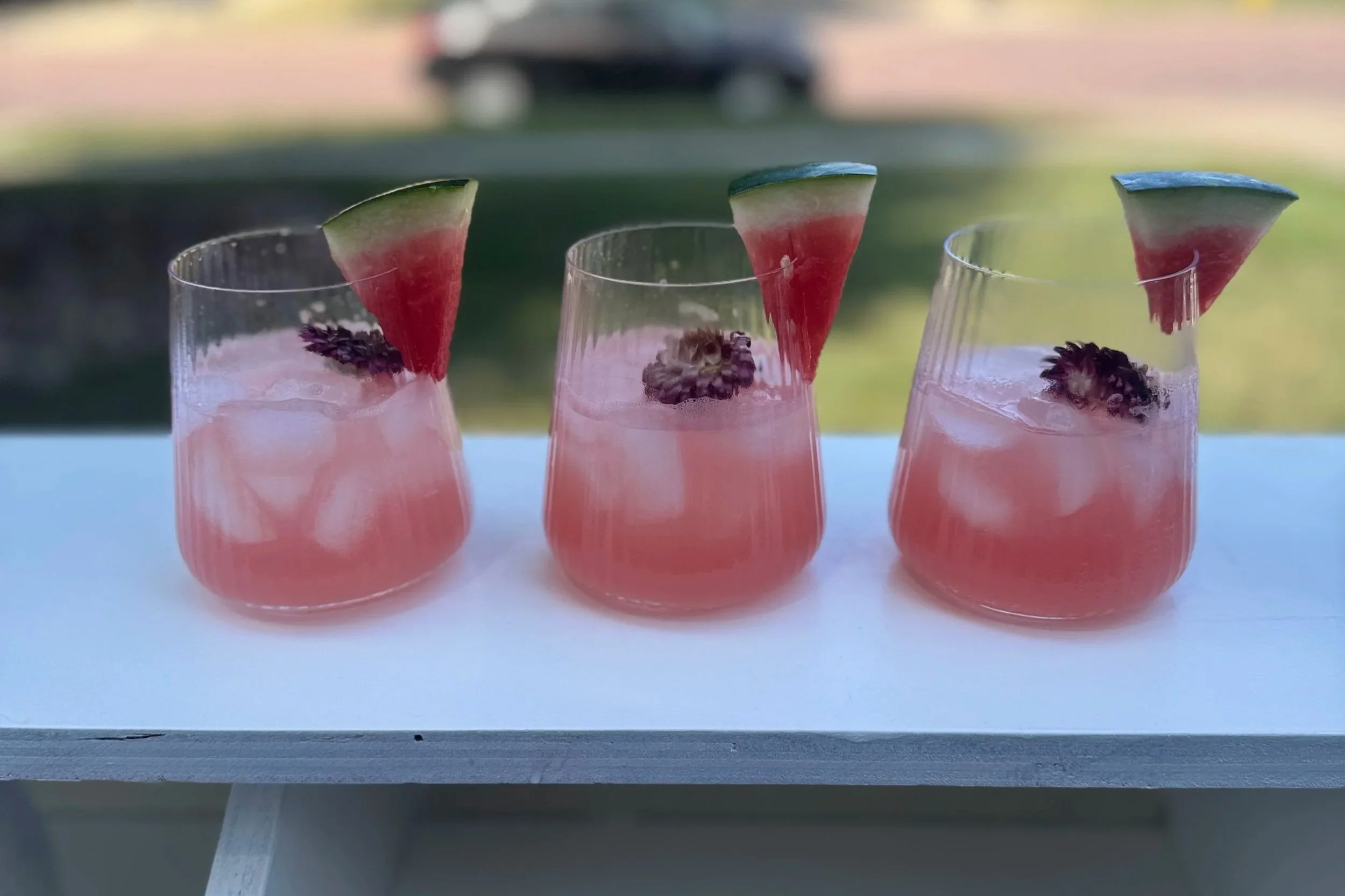 Three glasses of pink watermelon and blackberry cocktails garnished with small watermelon slices and blackberries, placed on a white surface outdoors.