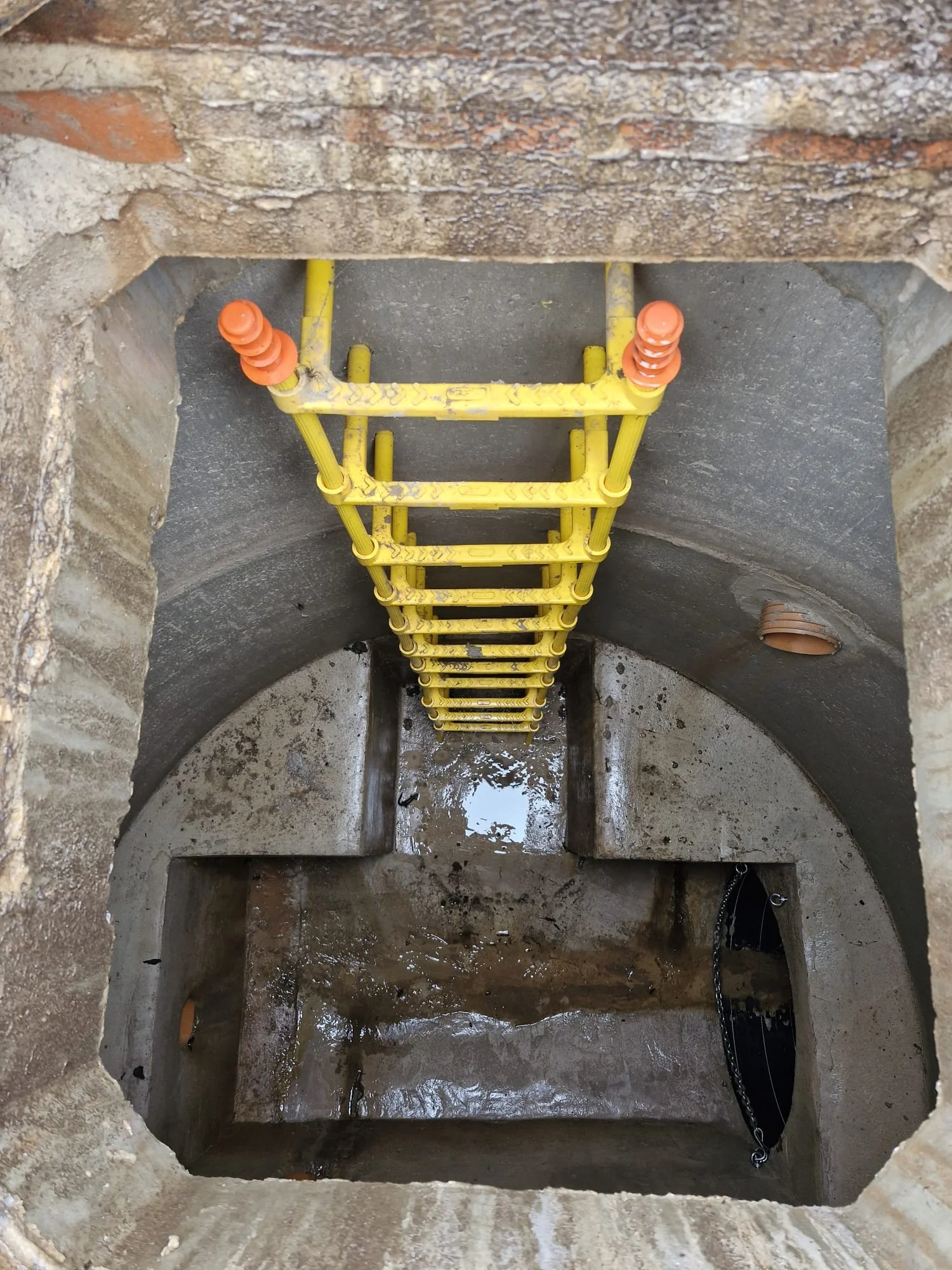 View of a construction site showing a concrete well with a yellow ladder leading down into water, surrounded by brick and concrete walls.