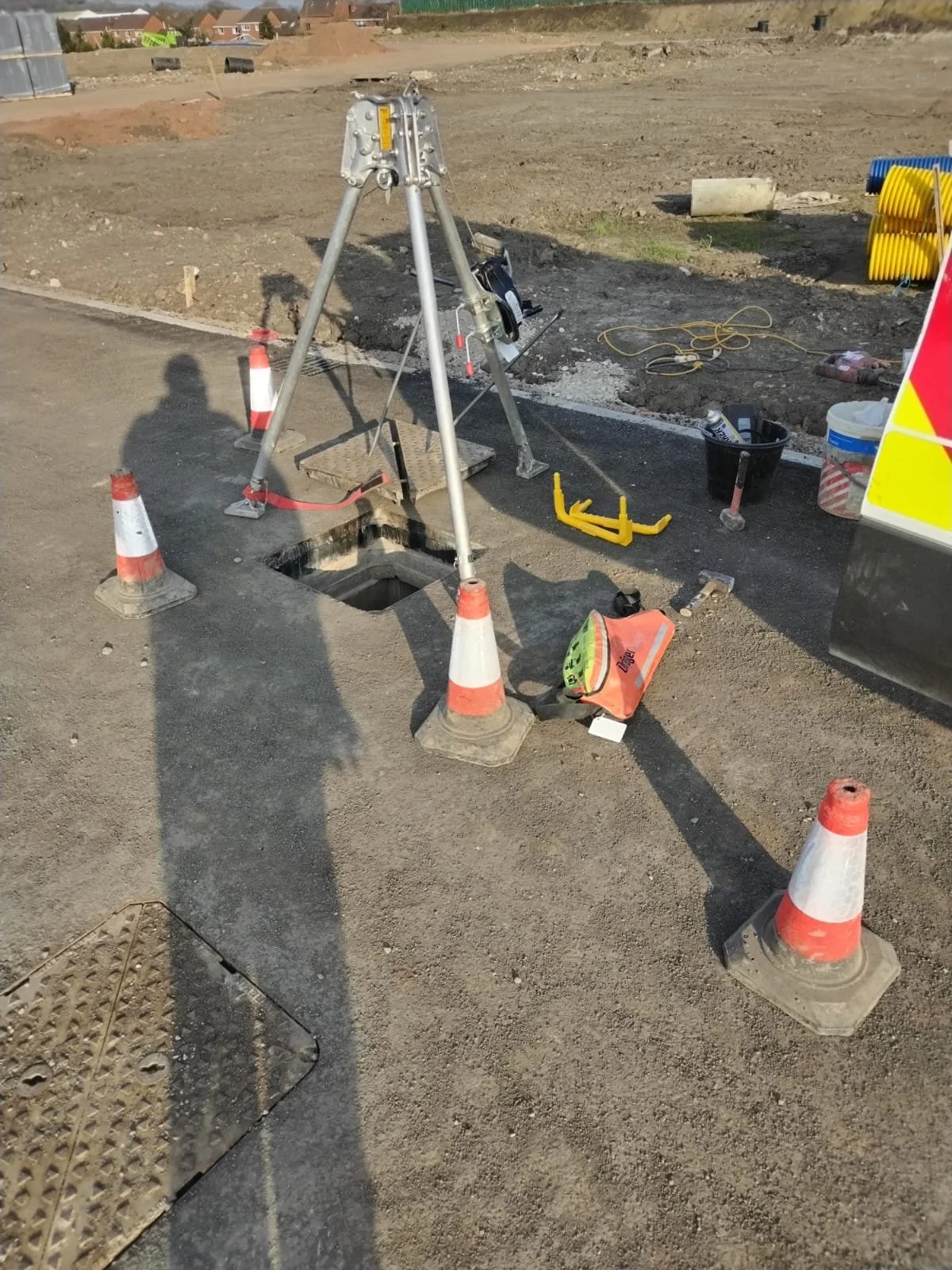 Construction site with orange and white traffic cones surrounding an open hole in the ground, various tools and equipment scattered around, and a tripod with surveying equipment set up near the hole.