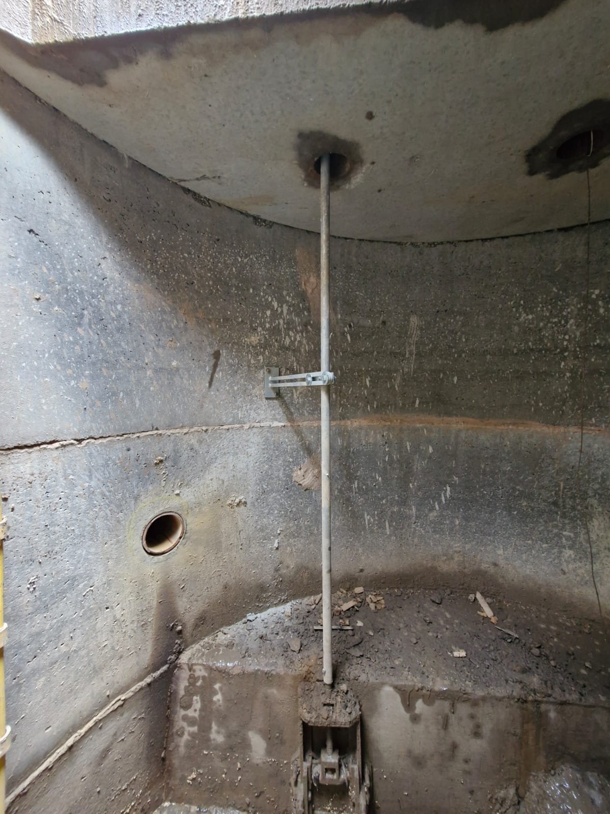 Underground utility access with pipes and conduit, rough concrete walls, and a vertical metal pipe in the center.