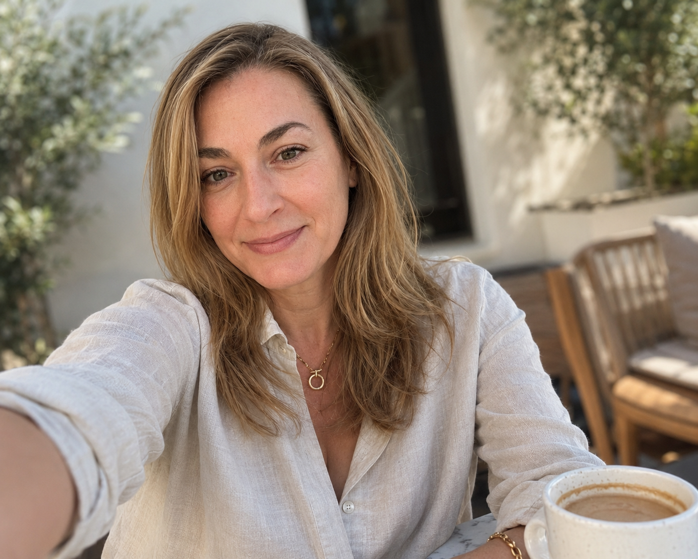 A woman with light brown hair and light skin, smiling and taking a selfie at an outdoor cafe, with a coffee mug in front of her.