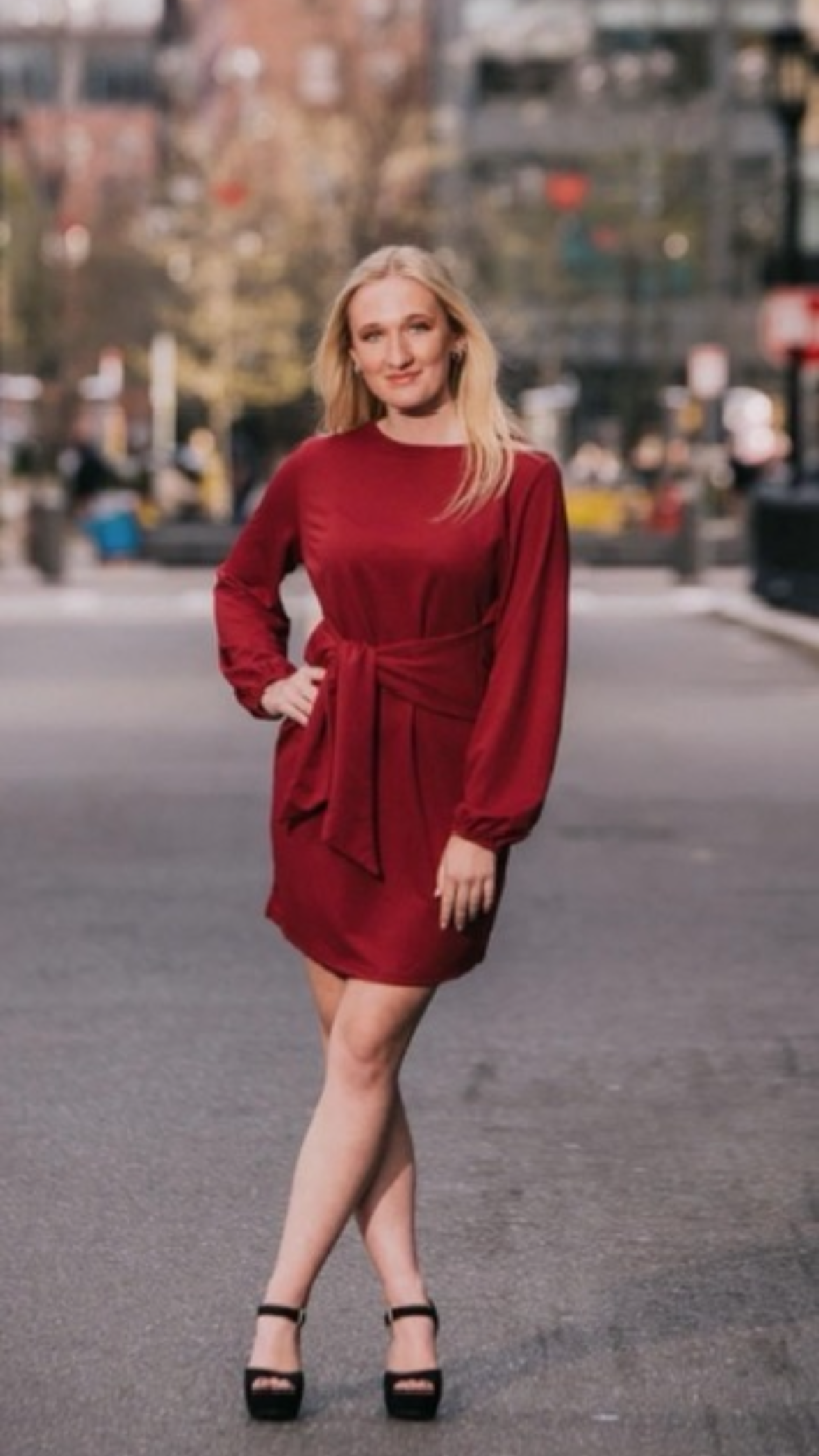A woman wearing a red dress and black high-heeled shoes standing on a city street with blurry buildings and trees in the background.