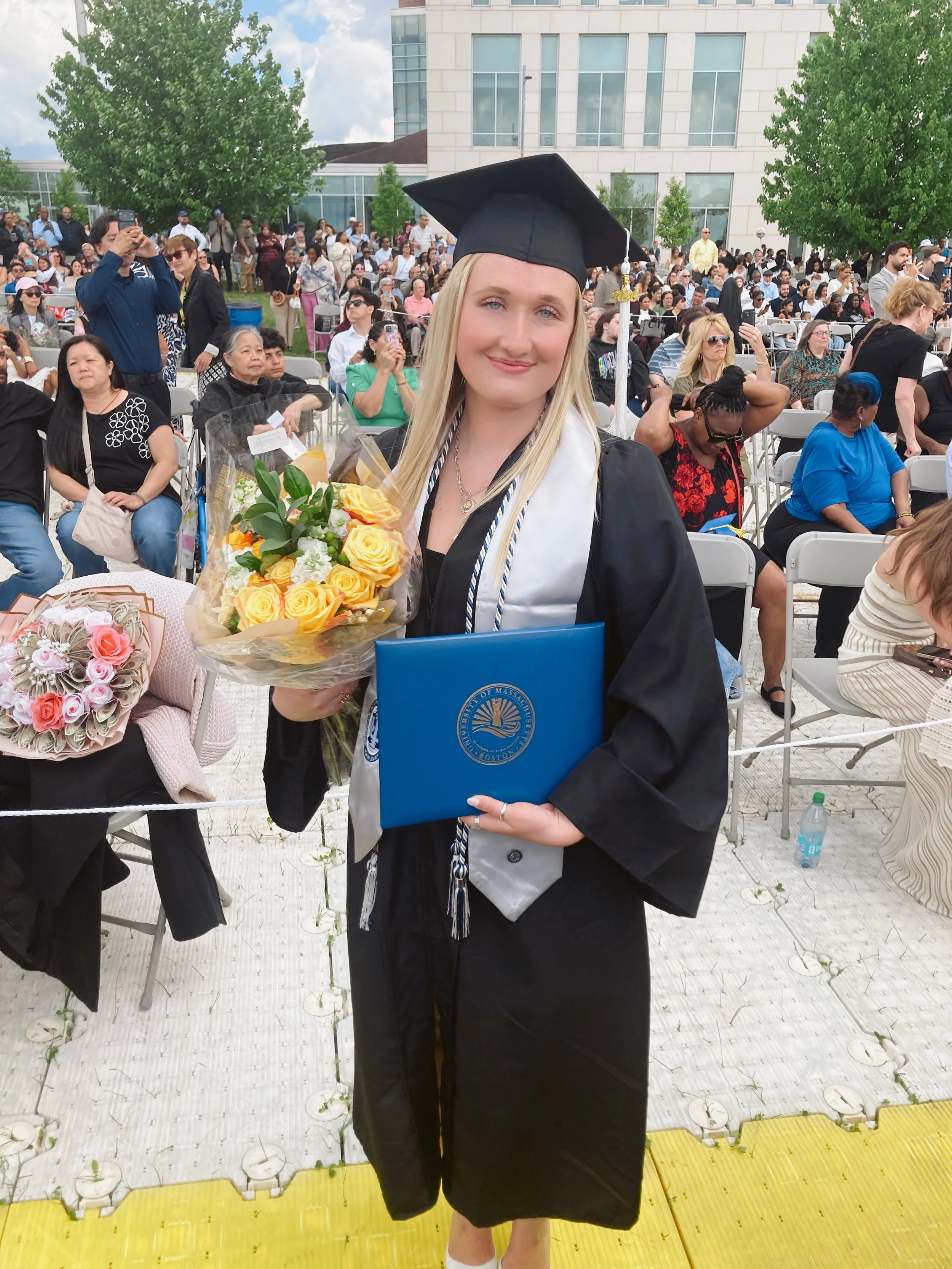 A young woman in a graduation cap and gown holding a bouquet of yellow roses and a blue diploma, smiling, at a graduation ceremony outdoor. Other people are seated and standing in the background.