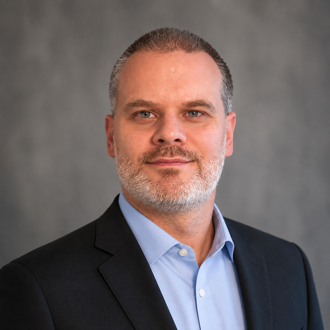 Professional headshot of a middle-aged man with grey hair and beard, wearing a black suit jacket and light blue dress shirt, standing against a grey background.