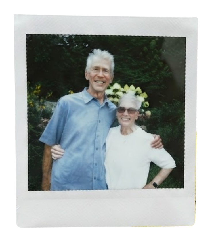 A smiling elderly man and woman standing outdoors, hugging each other. The man has white hair and is wearing a blue button-up shirt, while the woman has white hair, sunglasses, and is wearing a white shirt. Behind them, there are yellow and white flowers and green foliage.