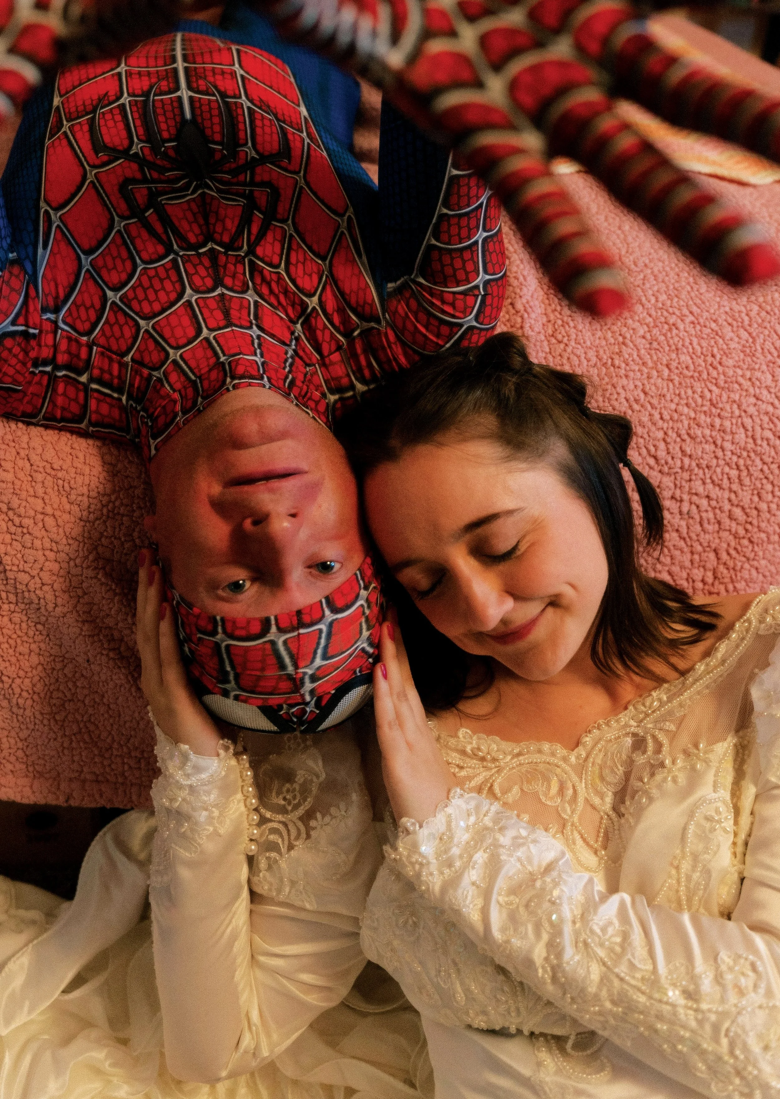 A woman in a cream-colored, lacy dress and a man in a Spider-Man costume lying down on a pink blanket, resting their heads together and smiling softly.