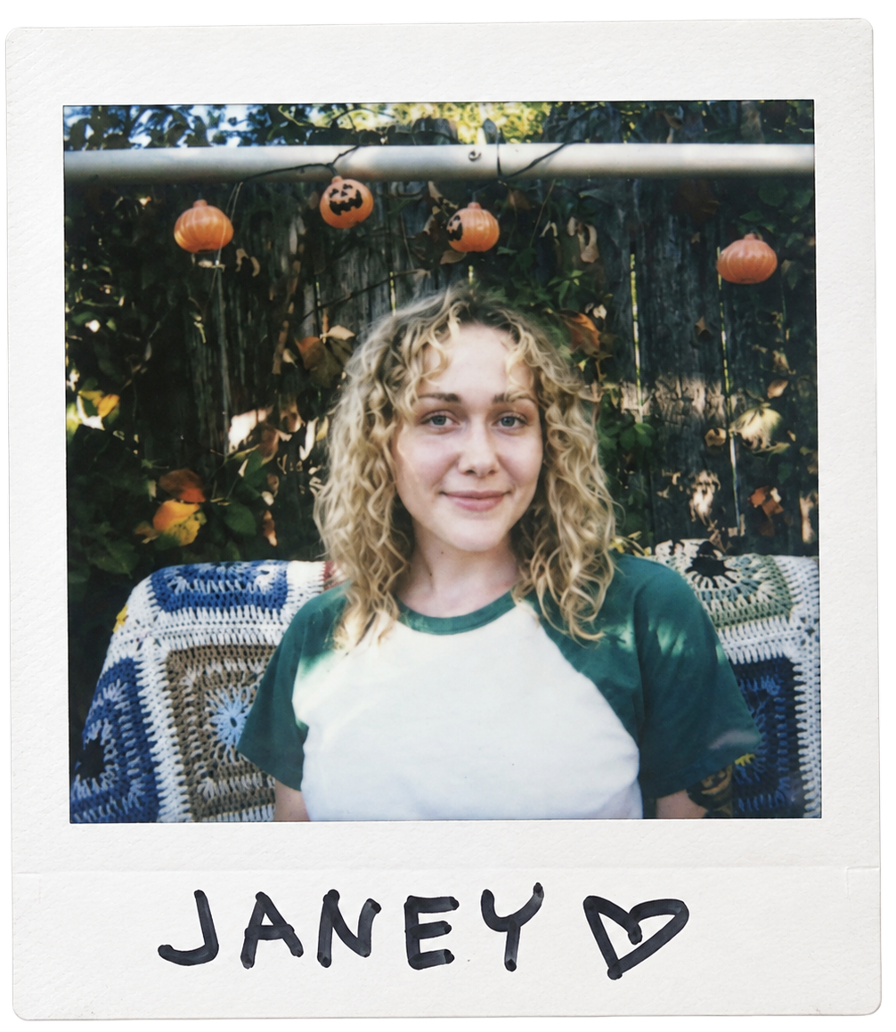 A young woman with curly blonde hair smiling outdoors in front of a garden fence adorned with small pumpkin lights. She is sitting on a crocheted blanket featuring blue, white, and beige patterns. The name 'JANEY' is handwritten at the bottom of the photo with a heart symbol.