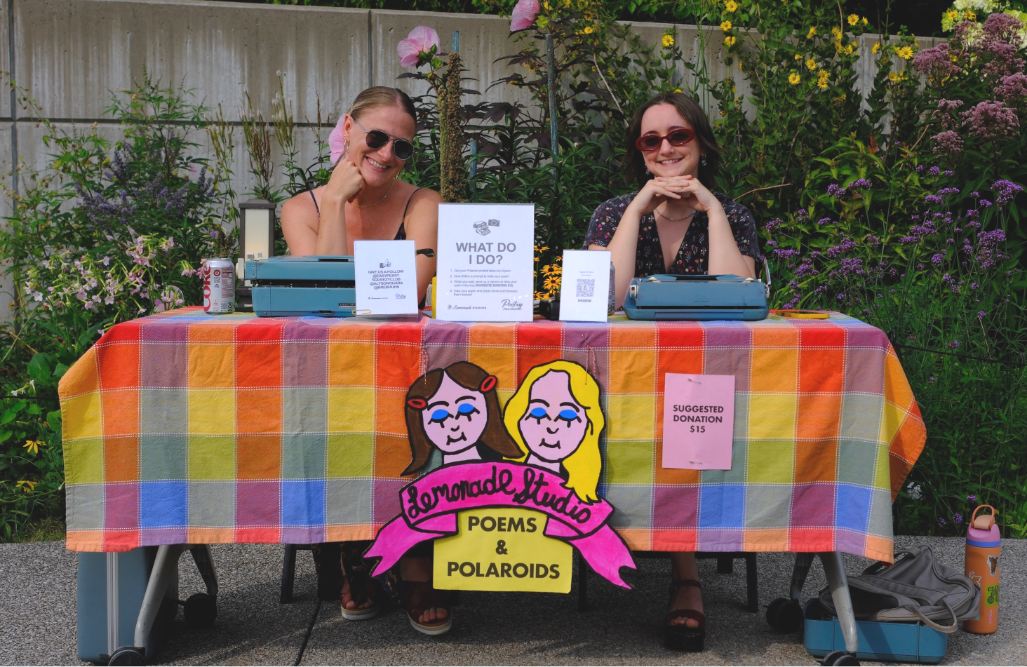 Two women sitting behind a rainbow-colored table at a poetry and polaroid event, smiling with typewriters, signs, and a cartoon sign with illustrated female faces, surrounded by flowers.