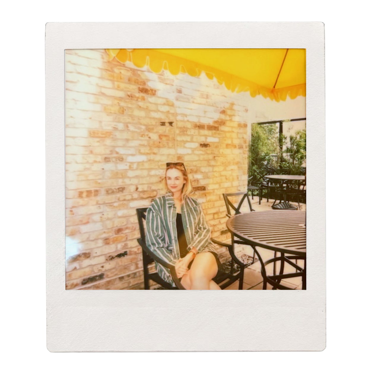 A woman sitting on a black chair at an outdoor patio with a brick wall behind her and a yellow canopy above. There is a round black table and other patio furniture around her.