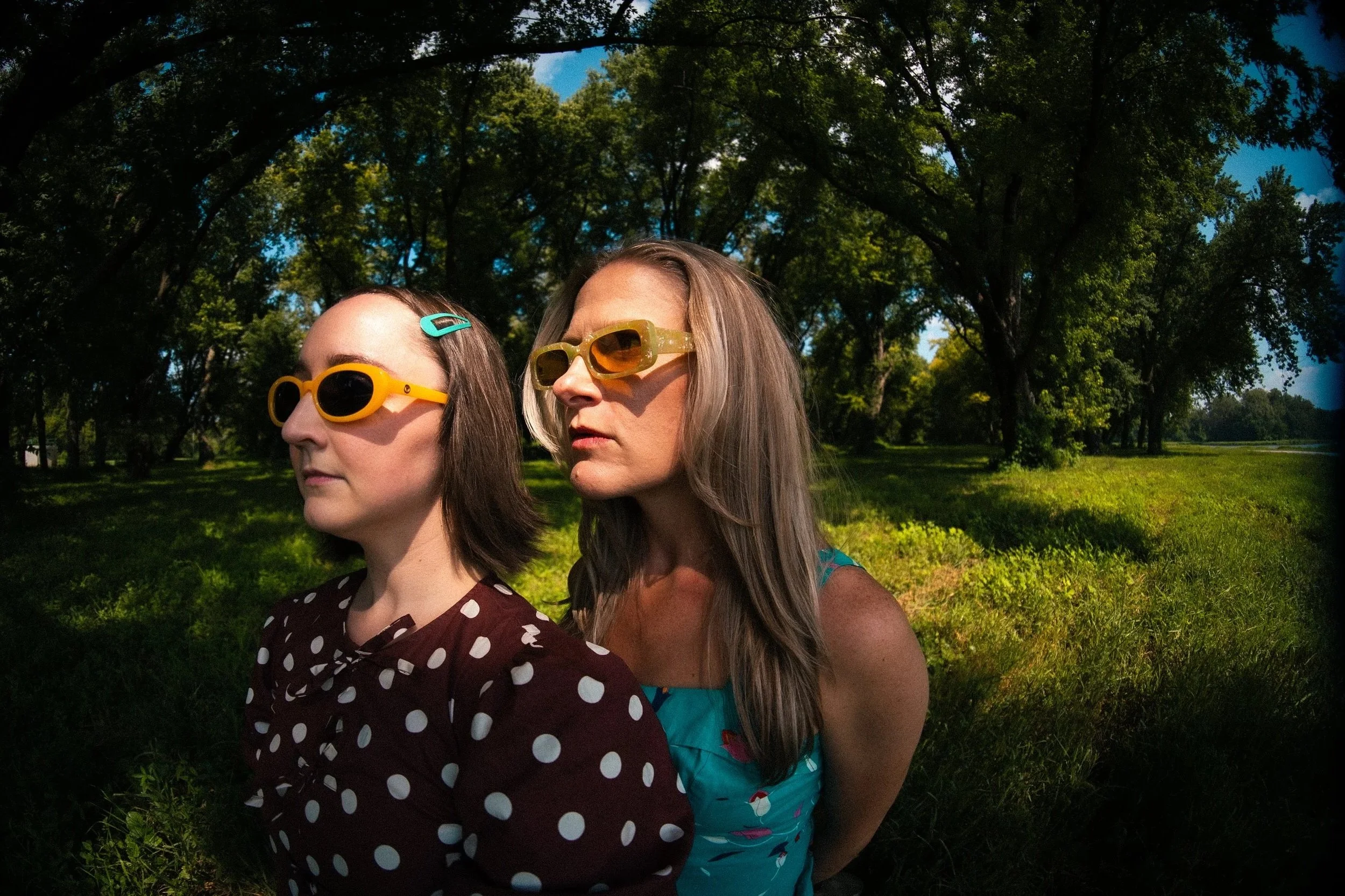Two women standing outdoors under trees on a sunny day, both wearing sunglasses. The woman on the left has dark hair, wearing dress with white polka dots, and a turquoise hair clip. The woman on the right has , wearing a turquoise dress with a floral pattern.