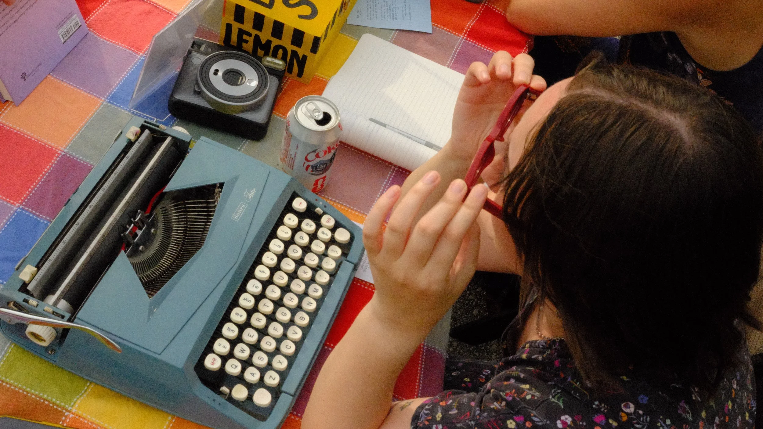 A young girl with brown hair and glasses is sitting at a table with a colorful tablecloth, holding her glasses and looking at a vintage blue typewriter surrounded by a soda can, a notebook, a camera, and some boxes.