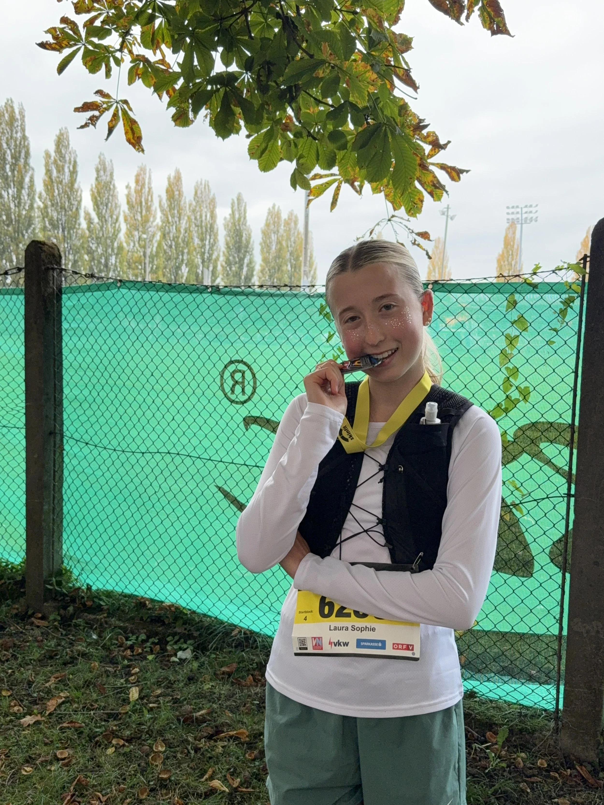 Eine junge Läuferin mit einer Startnummer 'Laura Sophie', eine Medaille um den Hals, steht vor einem grünen Zaun im Freien, umgeben von Bäumen und Laub auf dem Boden, während sie in einem Wettkampf lächelt .
