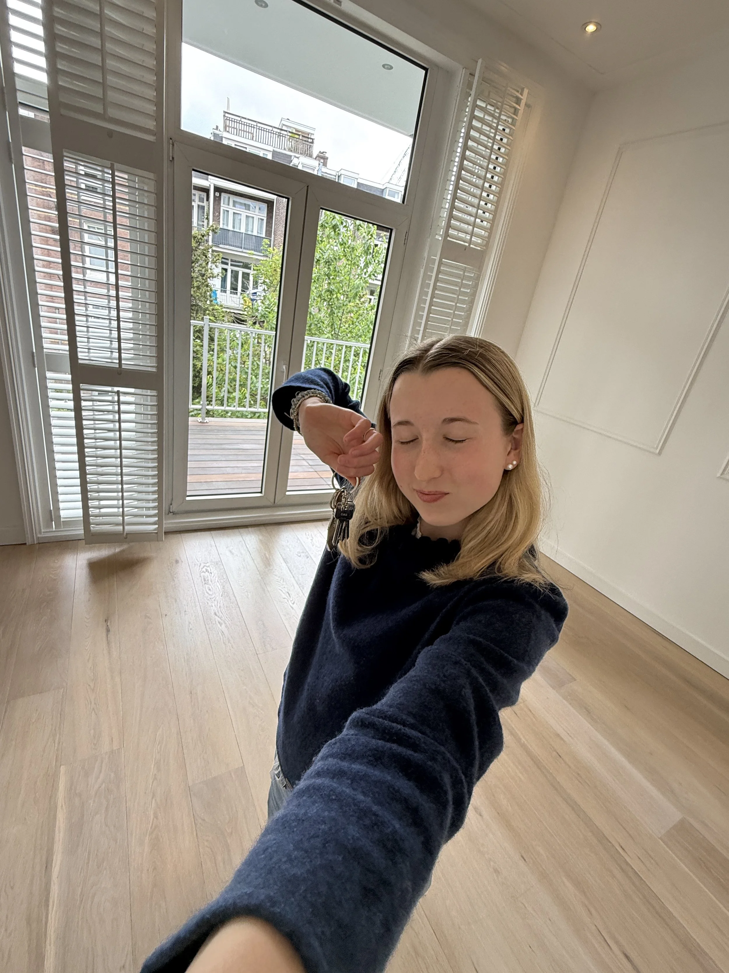 Ein junge Frau mit blonden Haaren macht ein Selfie in einem Raum mit Holzfußboden und großen Fenstern, die auf einen Balkon und ein Nachbarhaus mit Balkonen blicken. Ihre erste eigene Wohnung.