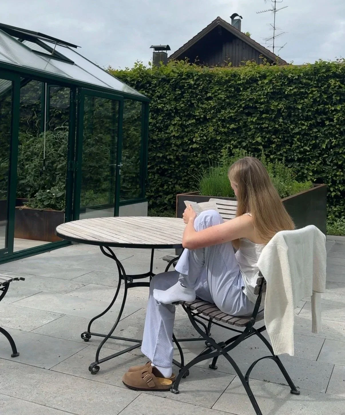 Eine junge Frau sitzt draußen auf einer Terrasse, liest ein Buch und hat eine weiße Jacke über die Rückenlehne ihres Stuhls gehängt. Im Hintergrund sind grüne Hecken, eine Glashaus und ein schwarzes Dach eines Hauses zu sehen.
