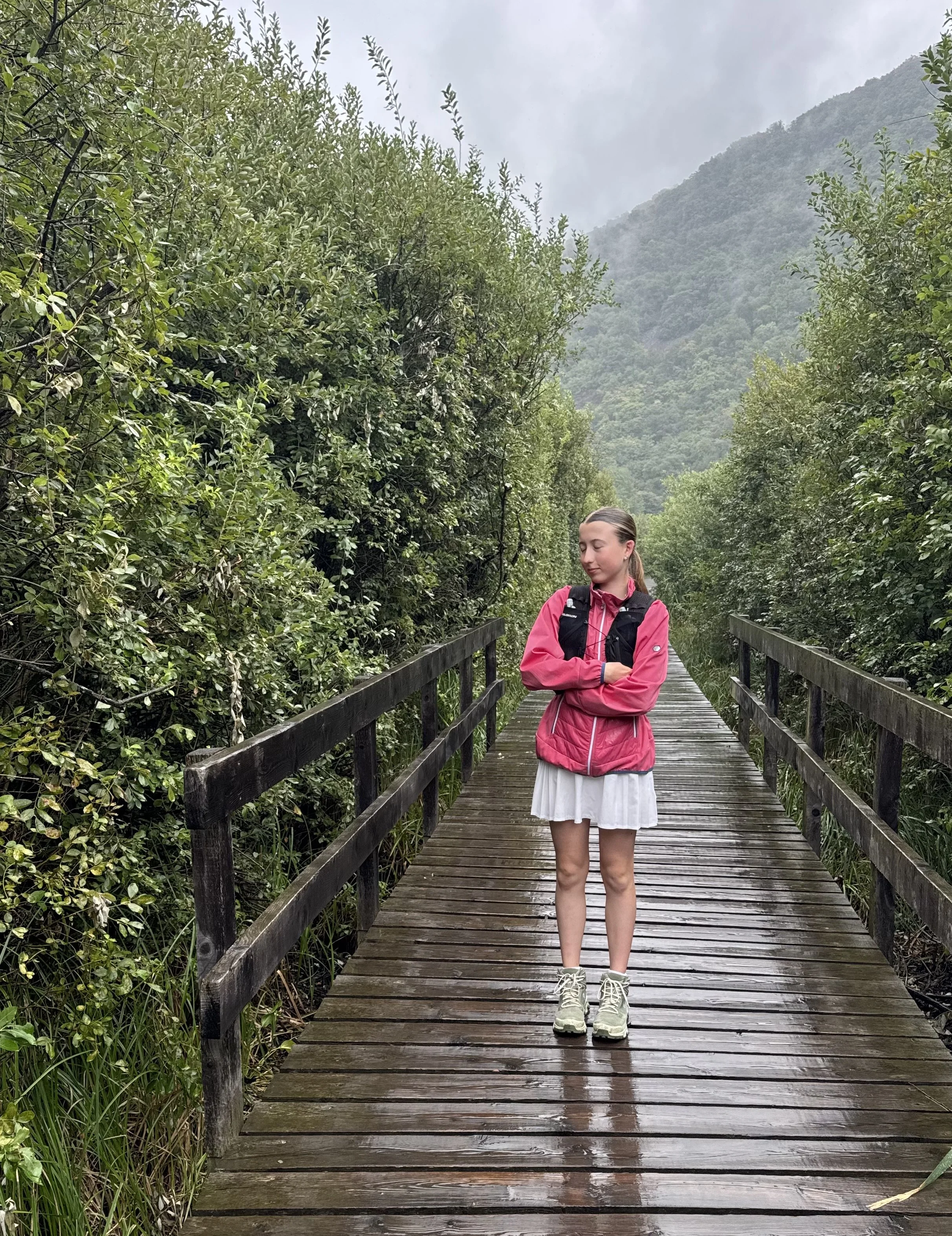 Eine junge Frau in einer pinken Jacke, weißen Tennisrock und Wanderschuhen steht auf einer hölzernen Brücke, umgeben von dichter grüner Natur und Bergen im Hintergrund, bei bewölktem Himmel.