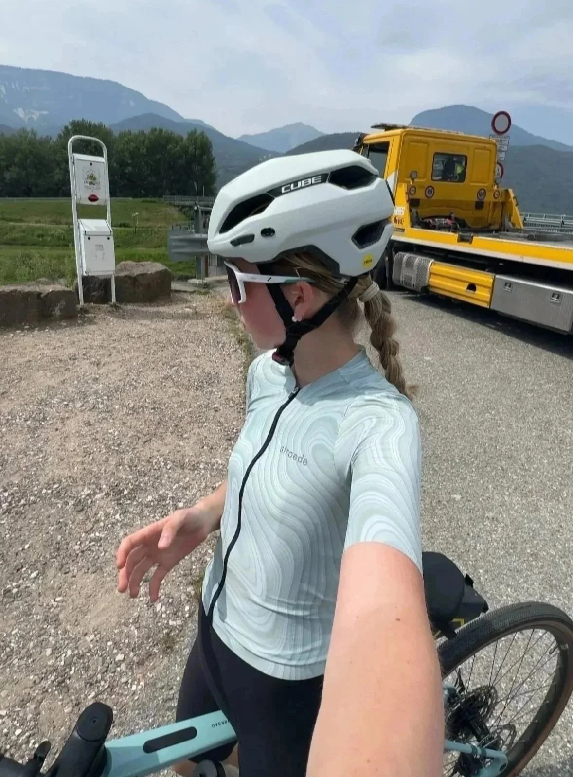 Junge Frau mit Fahrrad (Gravel Bike), weißer Fahrradhelm und Sonnenbrille, im Hintergrund gelber Abschleppwagen mit Bergen im Hintergrund.