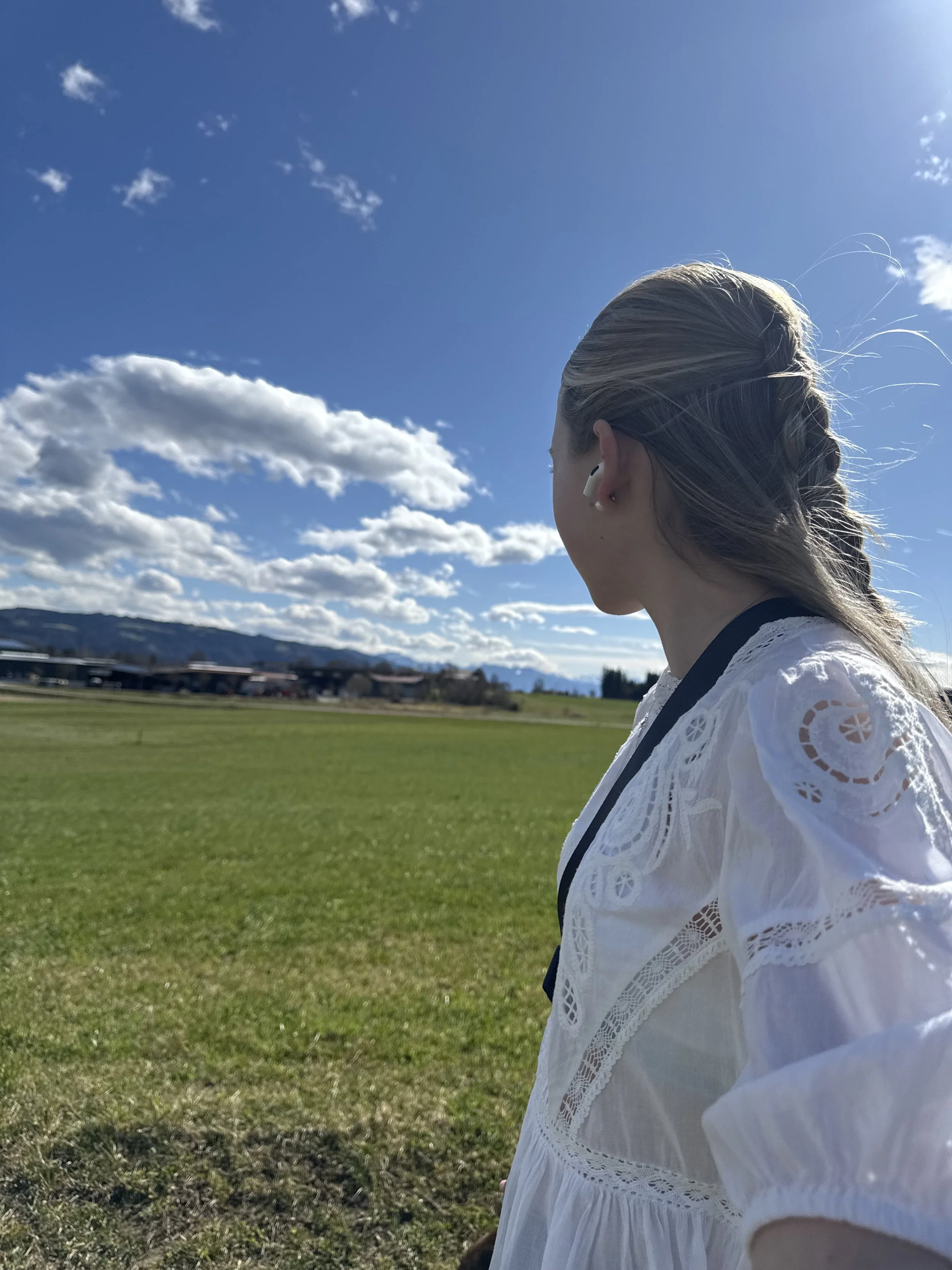 Seitenansicht einer jungen Frau mit geflochtenem Haar, weiße Spitzenbluse, Blick auf den blauen Himmel mit Wolken, in einer offenen grünen Landschaft bei sonnigem Wetter.