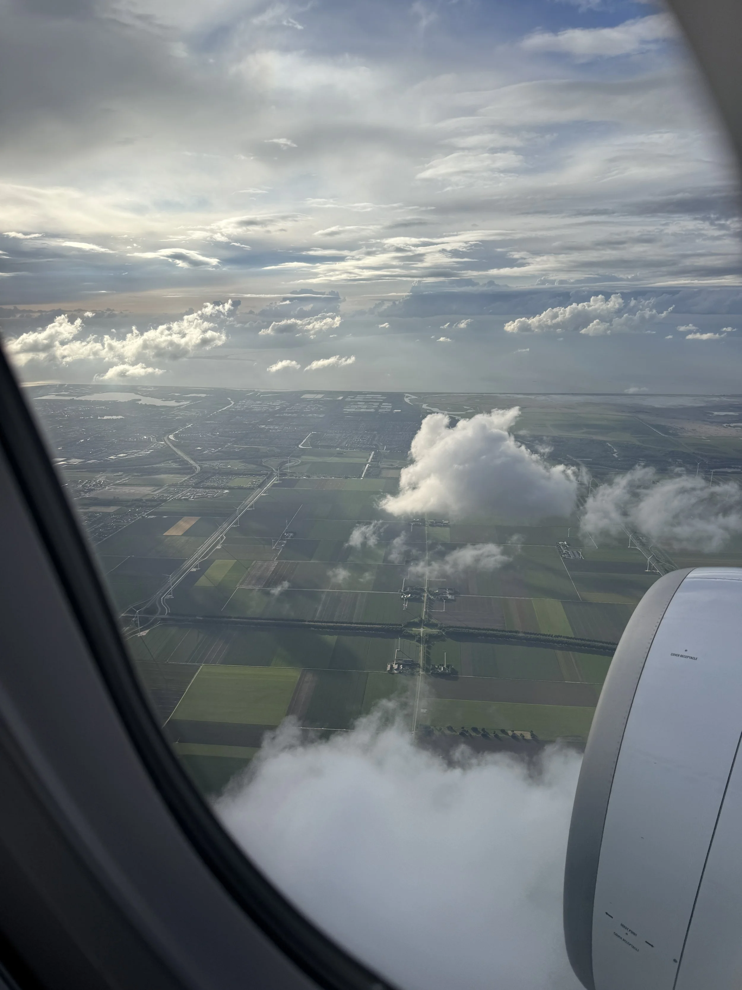 Der Blick aus einem Flugzeugfenster zeigt eine Landschaft mit Feldern, Straßen und Flüssen, unter einem bewölkten Himmel.