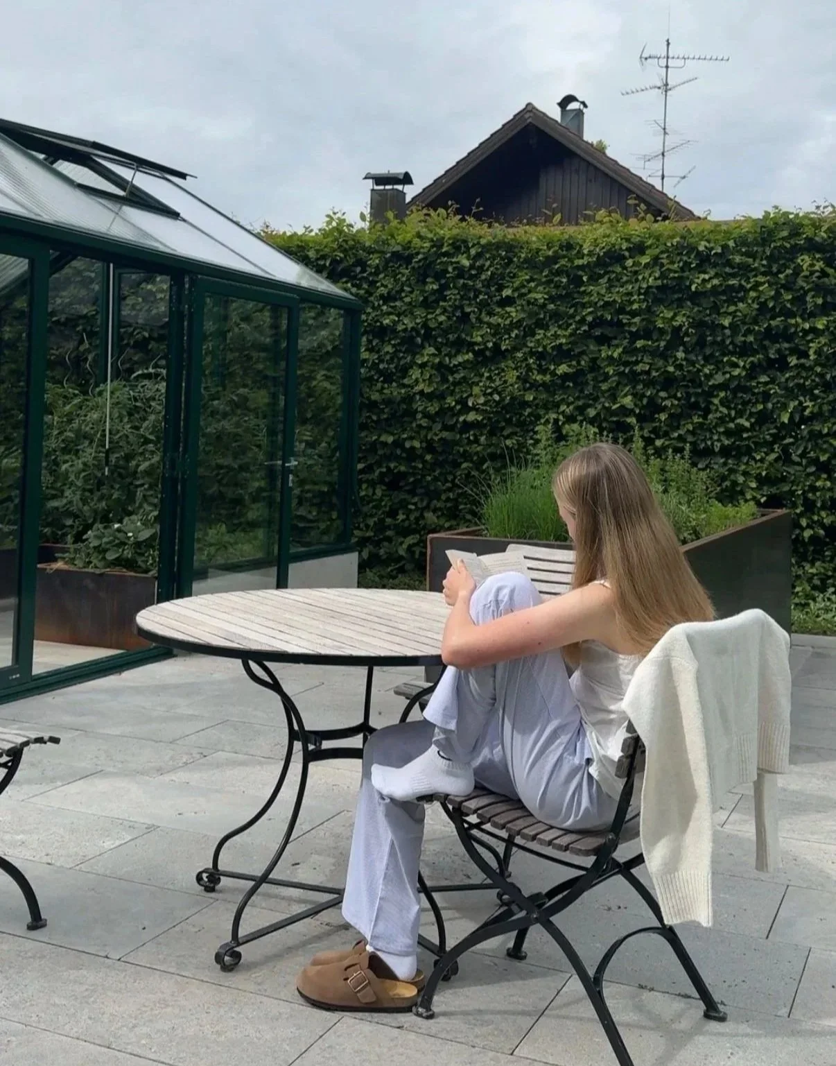 Junge Frau sitzt im Garten auf einem Stuhl, liest ein Buch, mit einem weißen Cardigan über der Rückenlehne. Im Hintergrund eine Hecke, ein Gewächshaus, und ein Haus mit mehreren Antennen auf dem Dach.