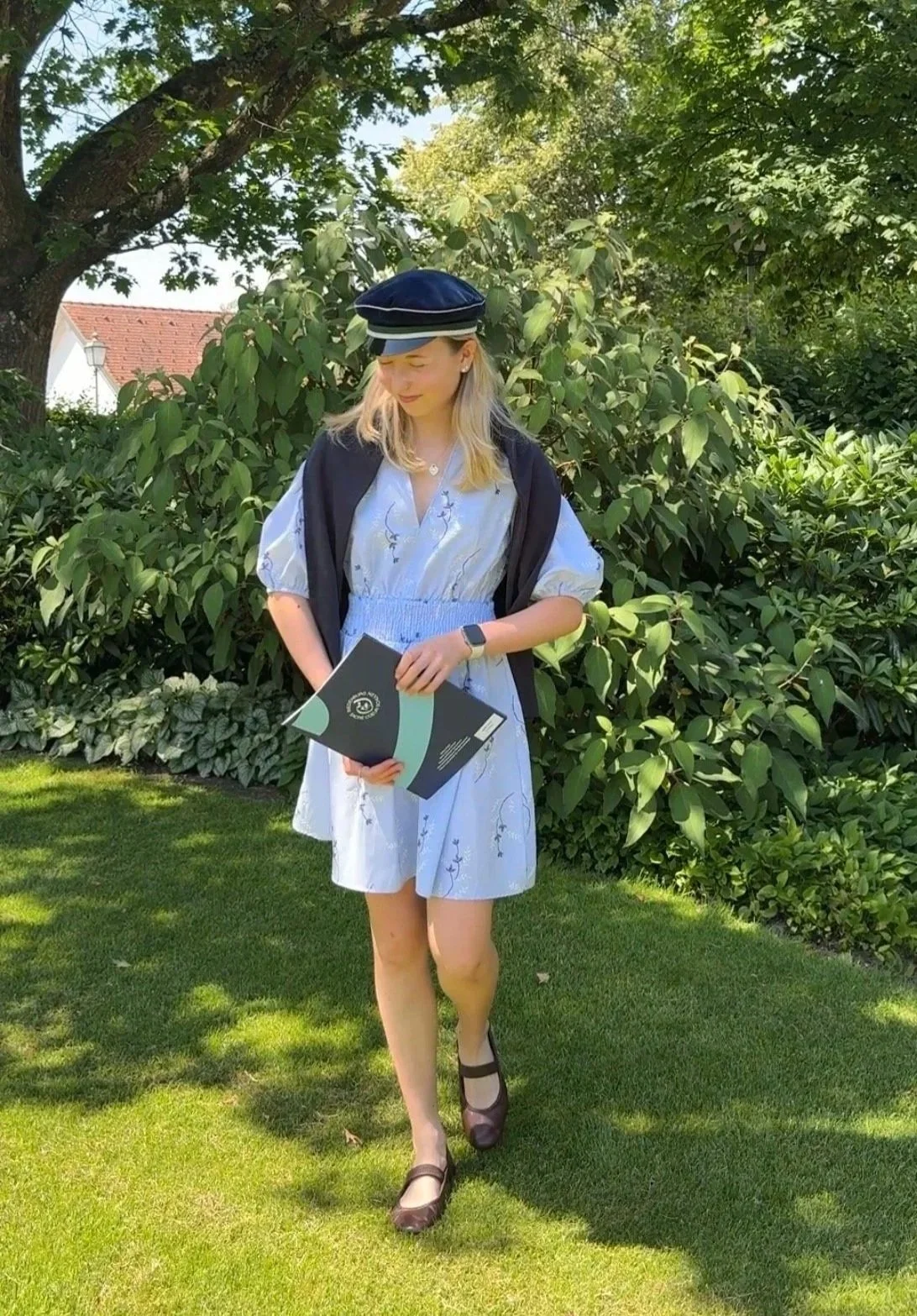 Junge Frau in Sommerkleid, mit braunen Ballerinas und dunkelblauem Käpple (Hut als Zeichen der Abschlussklasse an einem österreichischen Gymnasium), steht auf grünem Rasen vor Busch und Baum, hält ihr Zeugnis in der Hand, trägt eine Smartwatch.