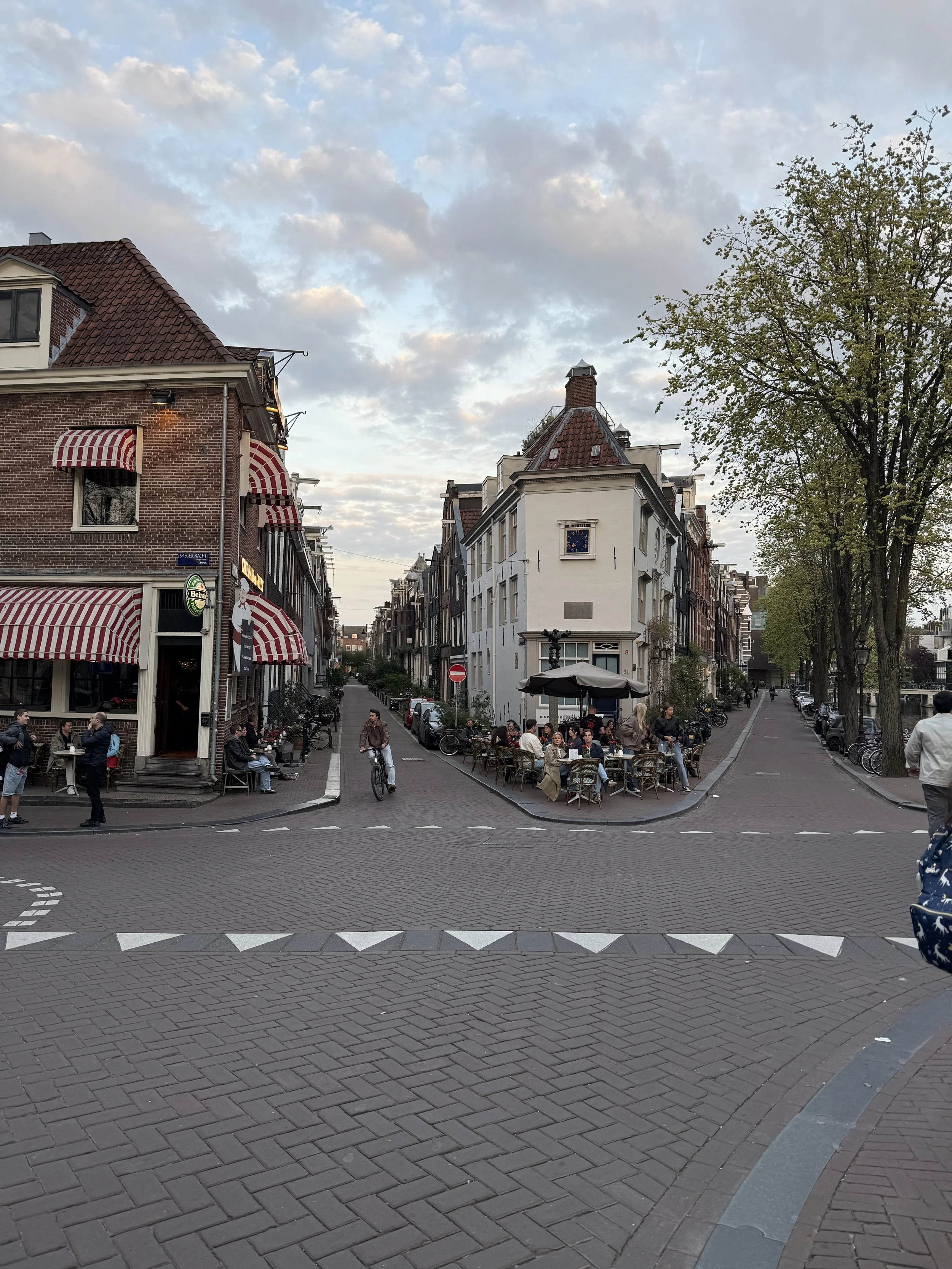 Straße in Amsterdam mit einigen Menschen, Tischen im Freien, alten Gebäuden, Bäumen und einem Blick in den Himmel mit Wolken.